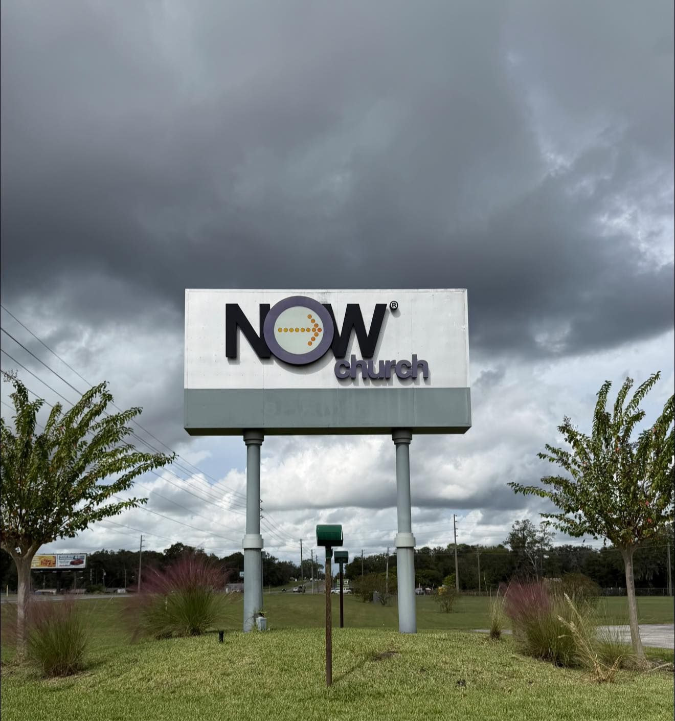 NOW Church sign on tall posts in a grassy lot under dark cloudy sky
