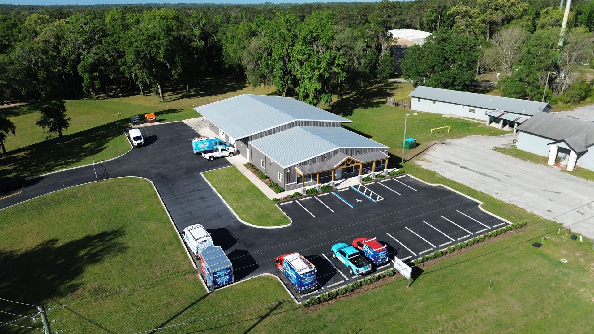Aerial view of a small building with a parking lot, several cars, and grassy grounds surrounded by trees