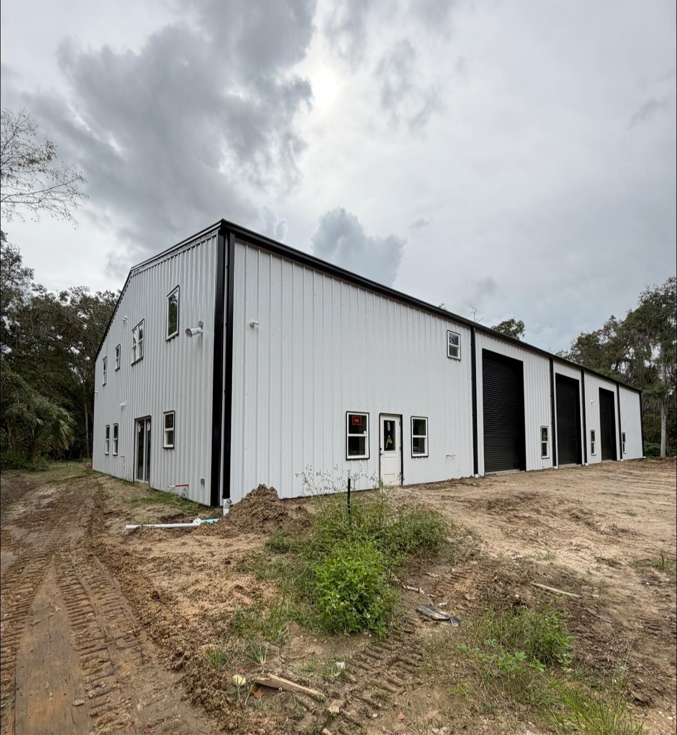 White metal barn-style building with black trim on a dirt lot under a cloudy sky