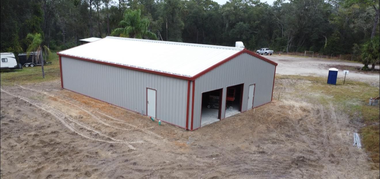 A large gray metal building with a red roof sits on a dirt lot near trees.