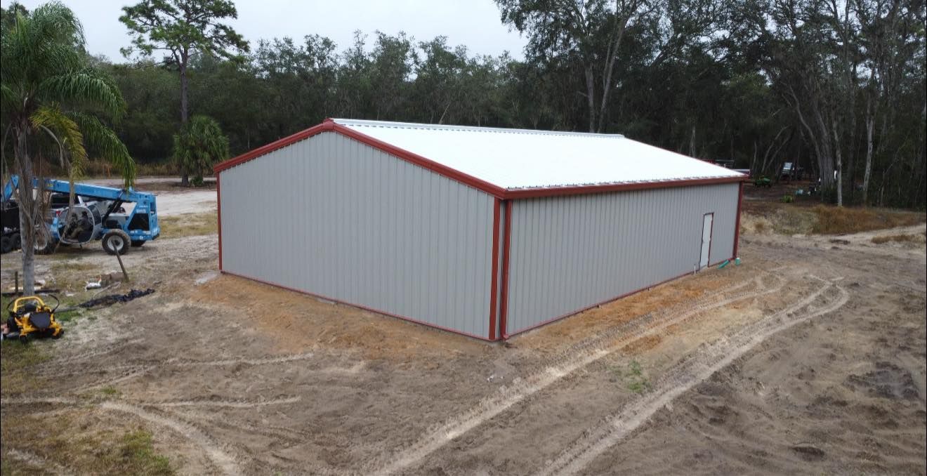 White metal storage building with red trim on a dirt lot, surrounded by trees and construction equipment.