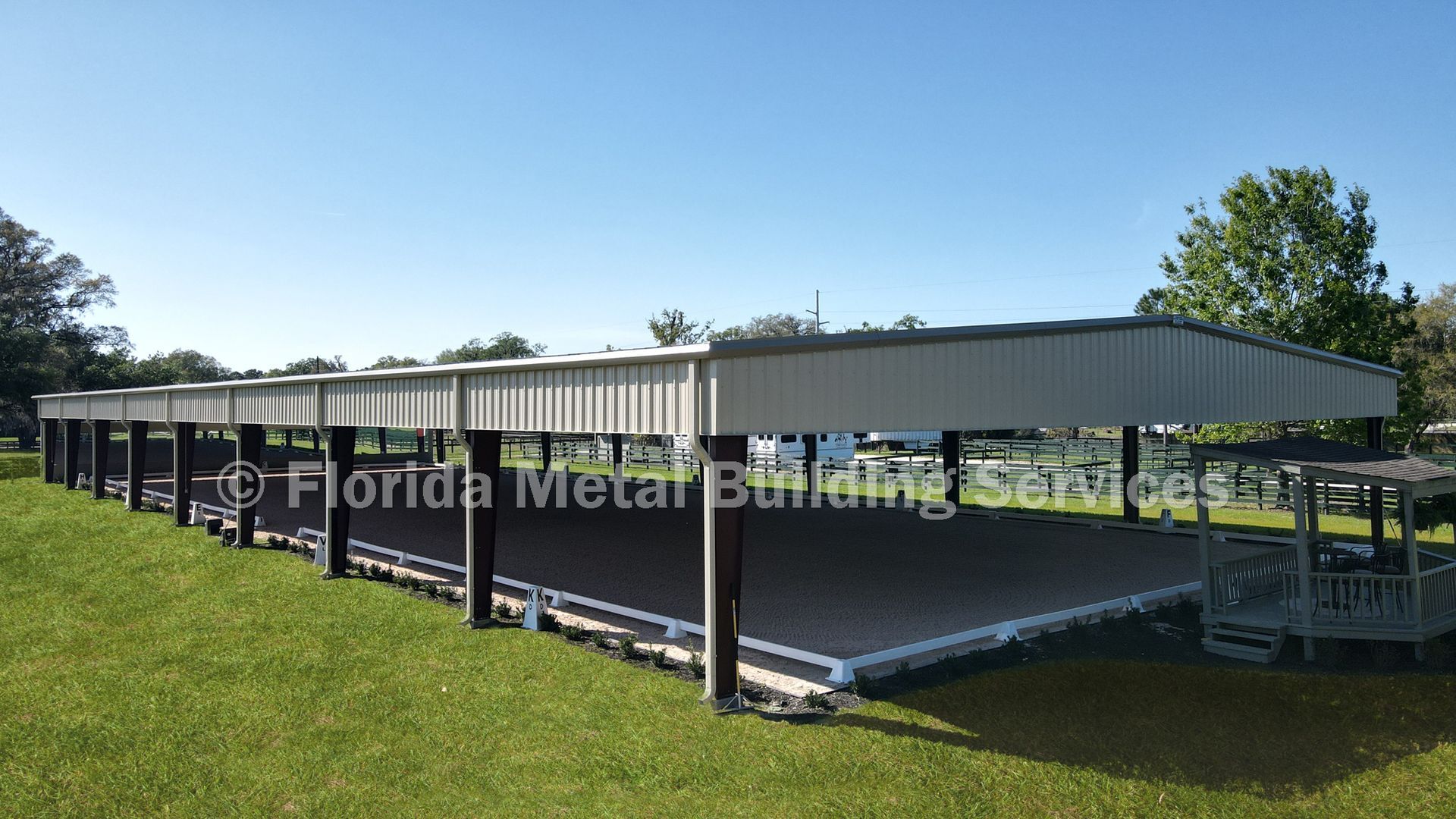 Long covered outdoor structure with black posts, metal roof, and grassy surroundings under a blue sky