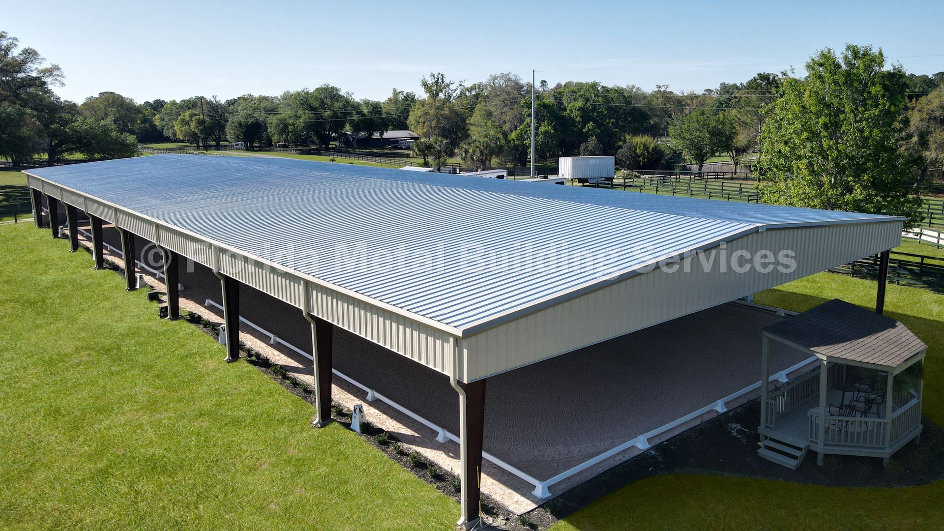Aerial view of a large metal-roofed pavilion with open sides, surrounded by grass and trees