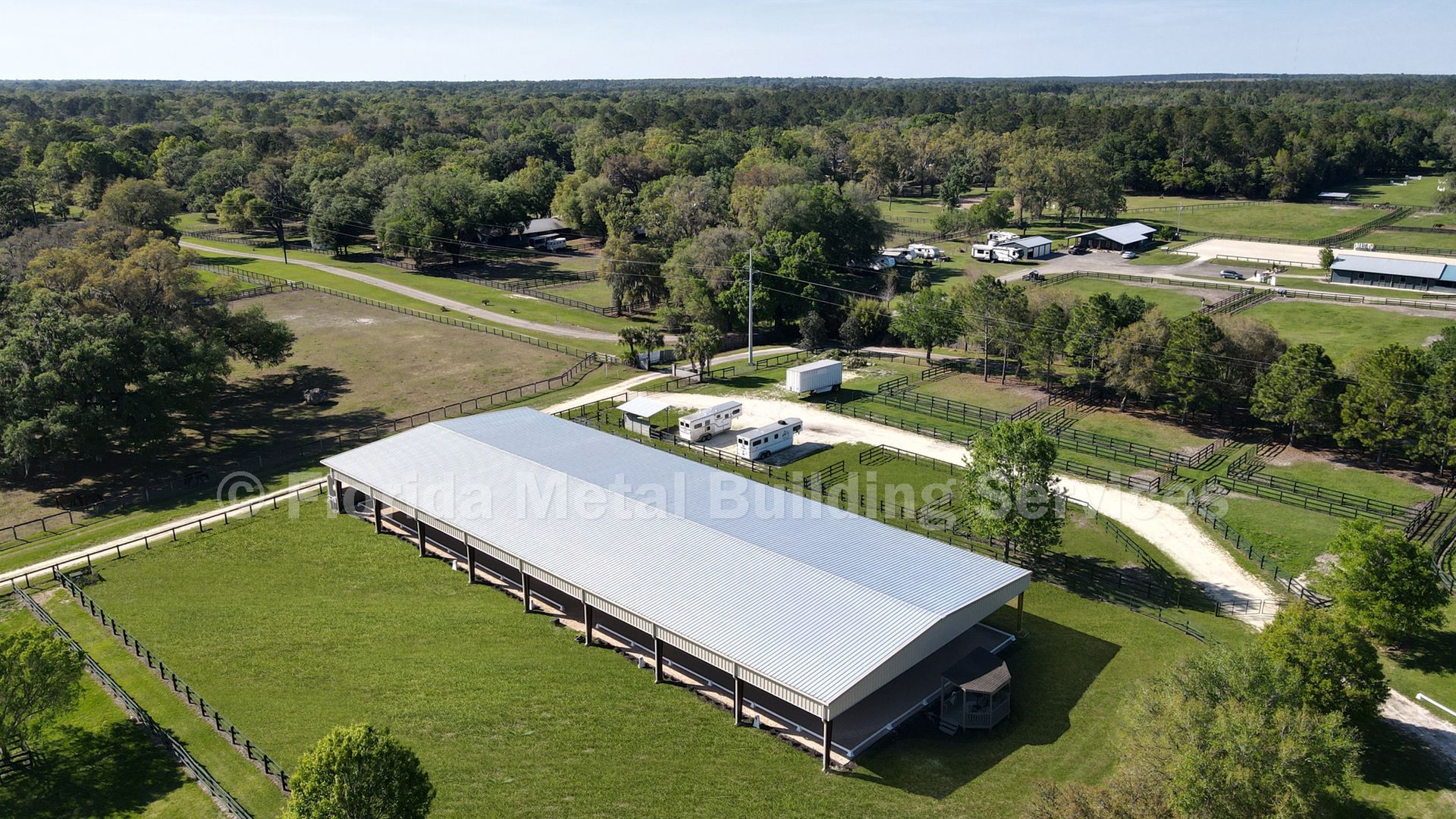Aerial view of a large white-roofed barn in a green rural landscape with trees and winding dirt roads.