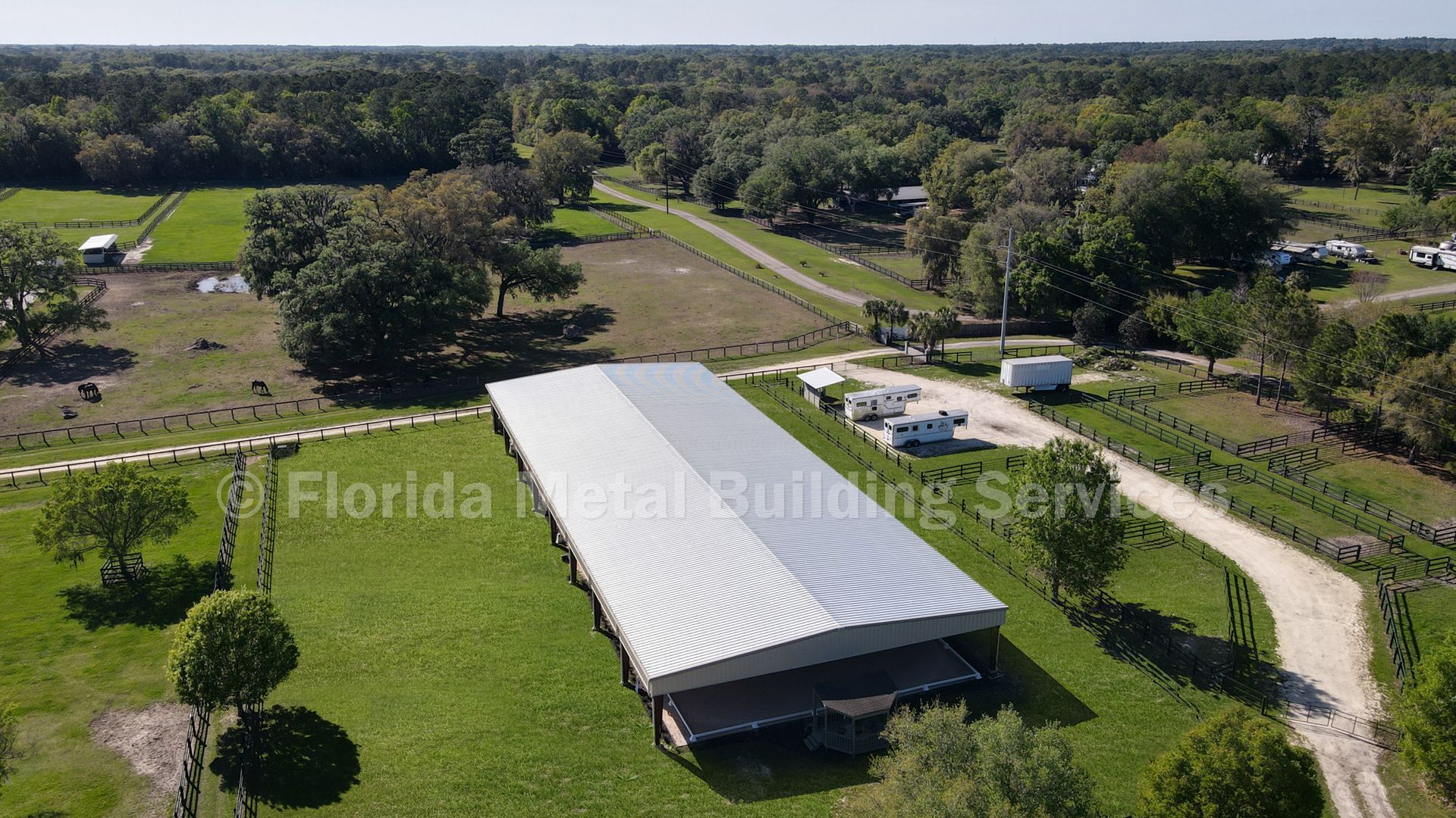 Aerial view of a long white-roofed barn on a grassy property with trees, dirt roads, and fields.