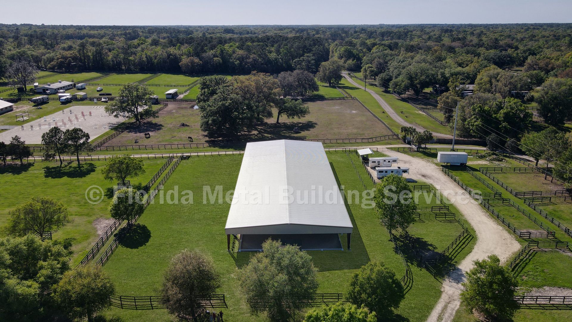 Aerial view of a white-roofed building surrounded by green fields, trees, and a winding dirt road.