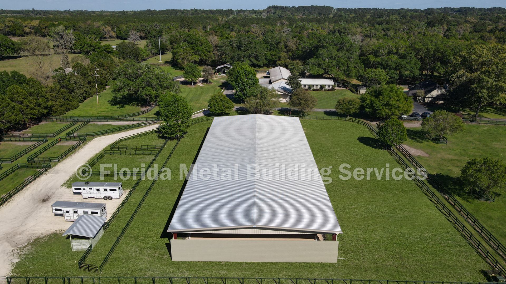 Aerial view of a large white metal-roof building with a striped pattern, surrounded by trees and grassy lots