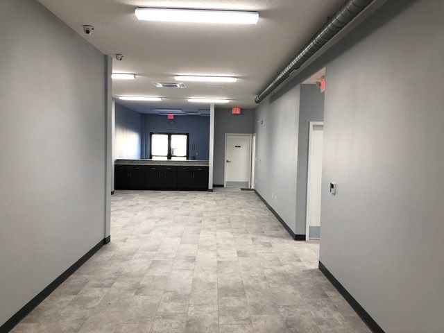 Empty office hallway with gray walls, carpet tiles, and fluorescent ceiling lights leading to a reception desk.