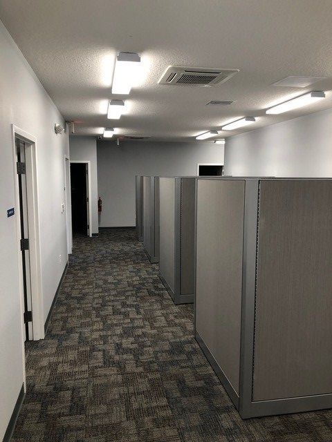 Empty office hallway with gray cubicle partitions, patterned carpet, and ceiling lights