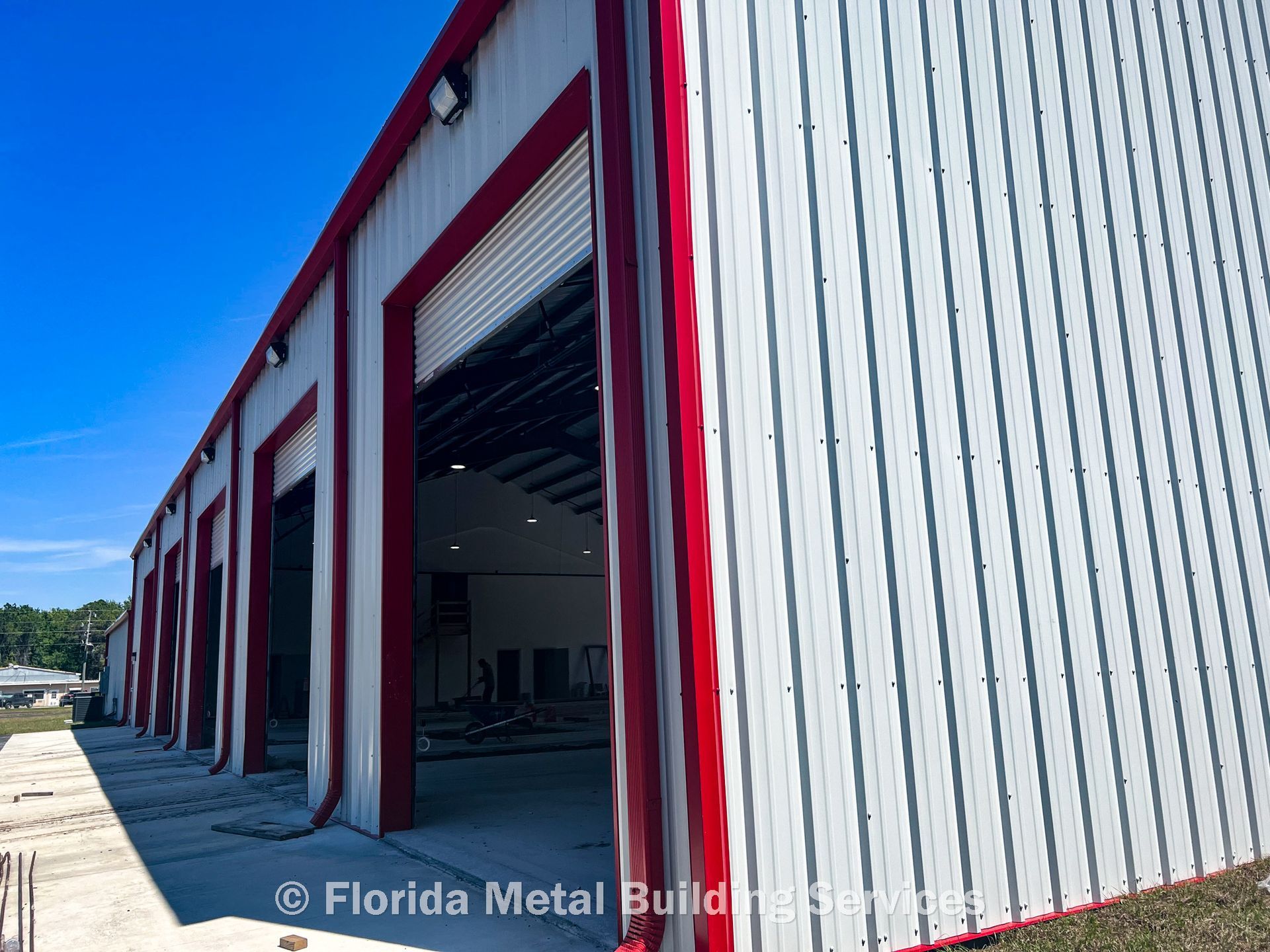 A side view of a large metal building with white vertical siding, red trim, and several open bay doors under a blue sky.
