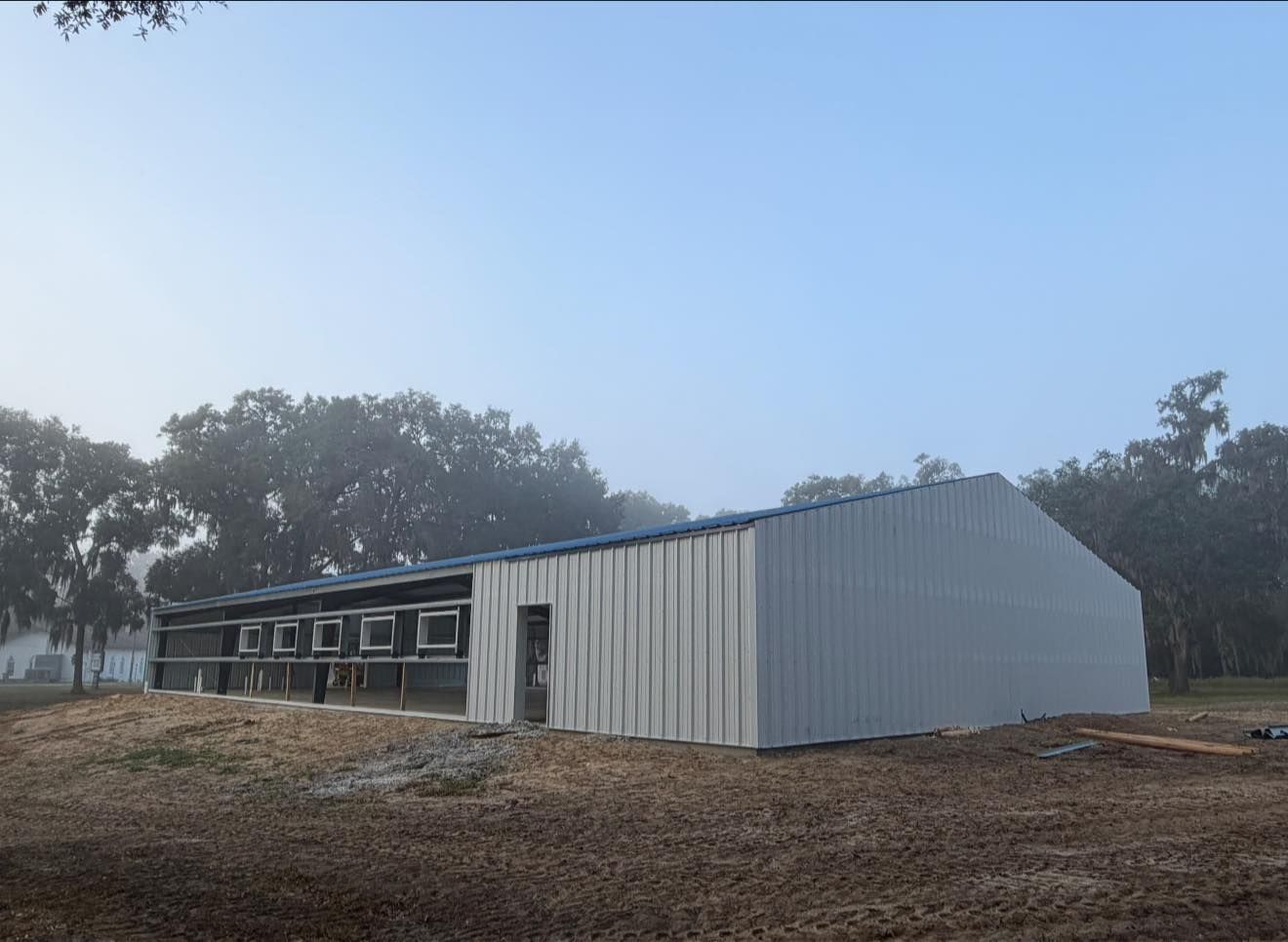 A white, metal-sided agricultural building stands on a dirt lot, surrounded by trees under a misty, pale blue sky.