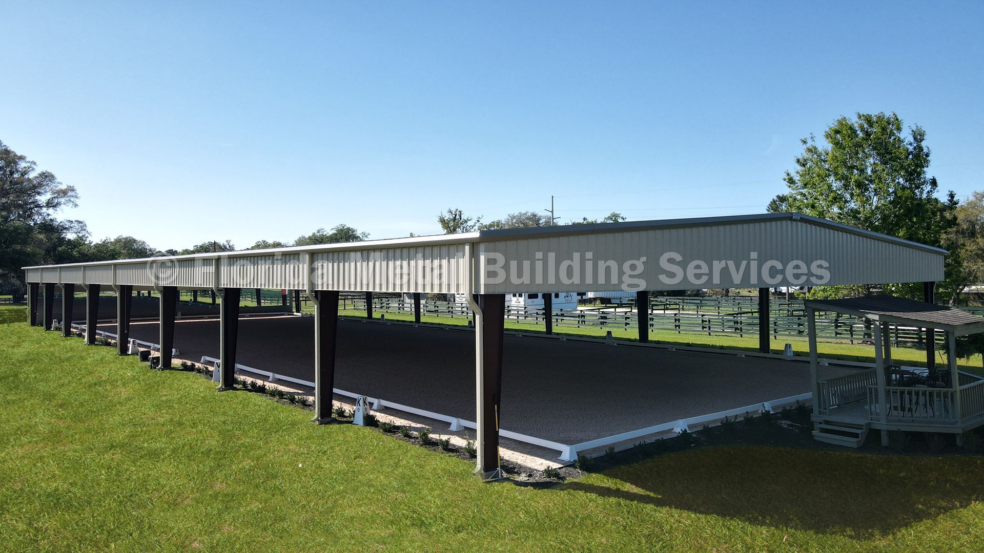 A large, rectangular, open-sided metal structure covering a horse riding arena in a grassy field.