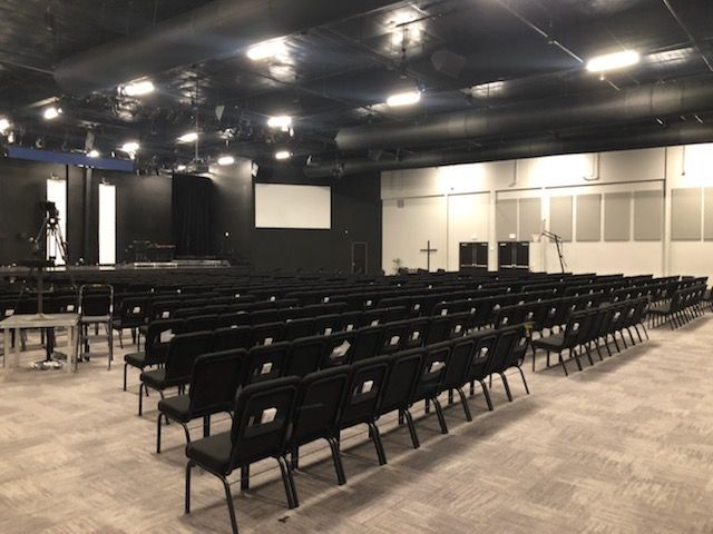 Empty auditorium with rows of black chairs, stage, and projector screen under bright ceiling lights