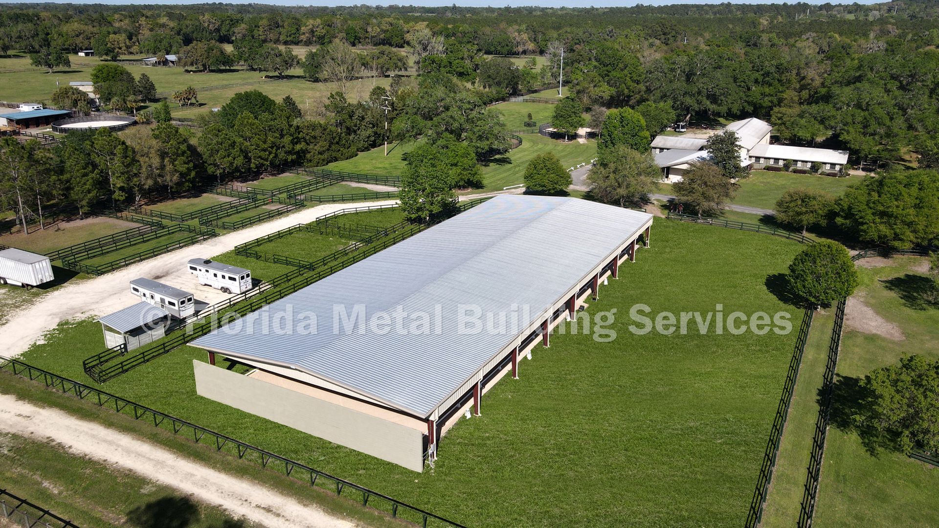 Aerial view of a long, metal-roofed barn structure surrounded by green fields and trees on a rural property.