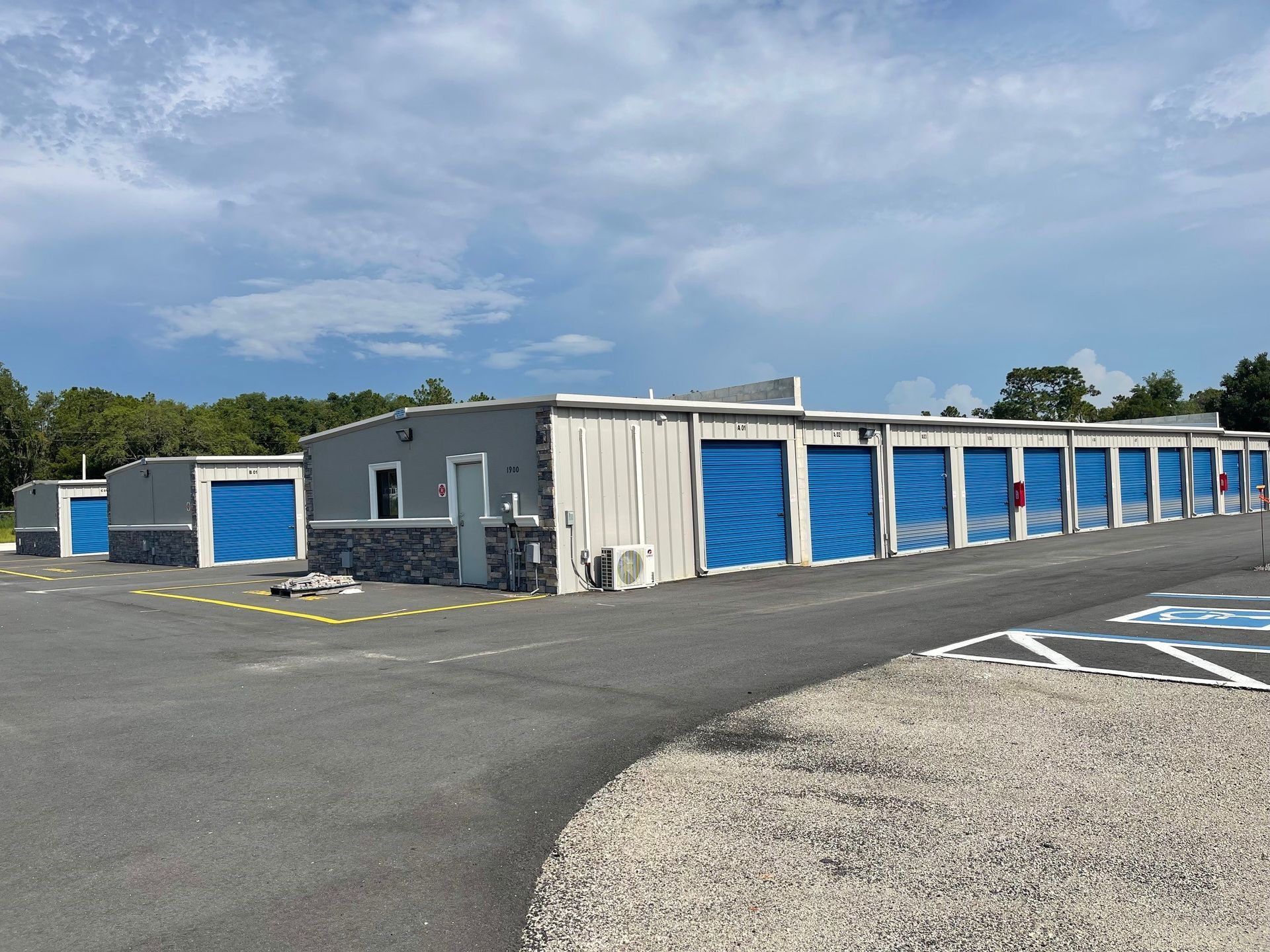 A long, single-story gray self-storage facility with multiple blue roll-up doors, set against a blue, cloudy sky.