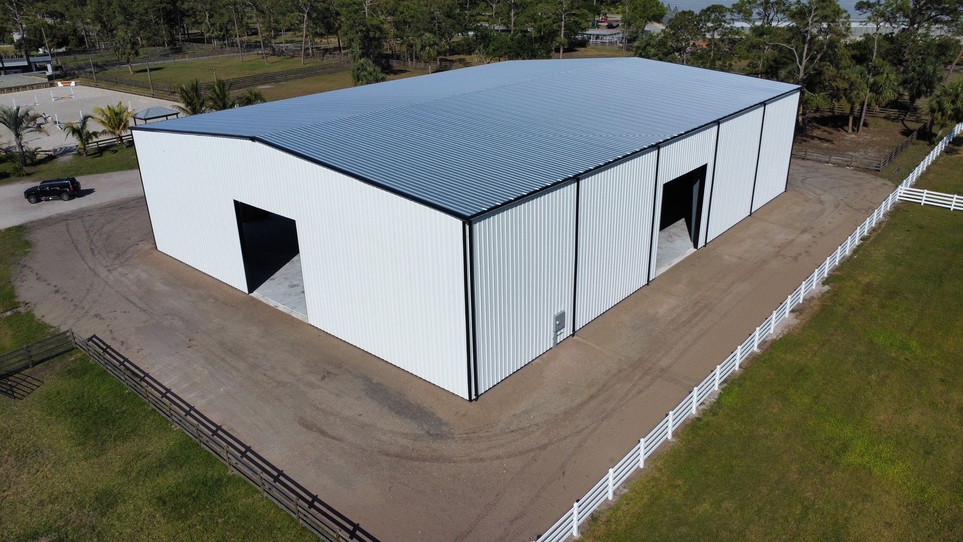 Aerial view of a white, rectangular warehouse building with a metal roof, surrounded by a white fence and dirt driveway.