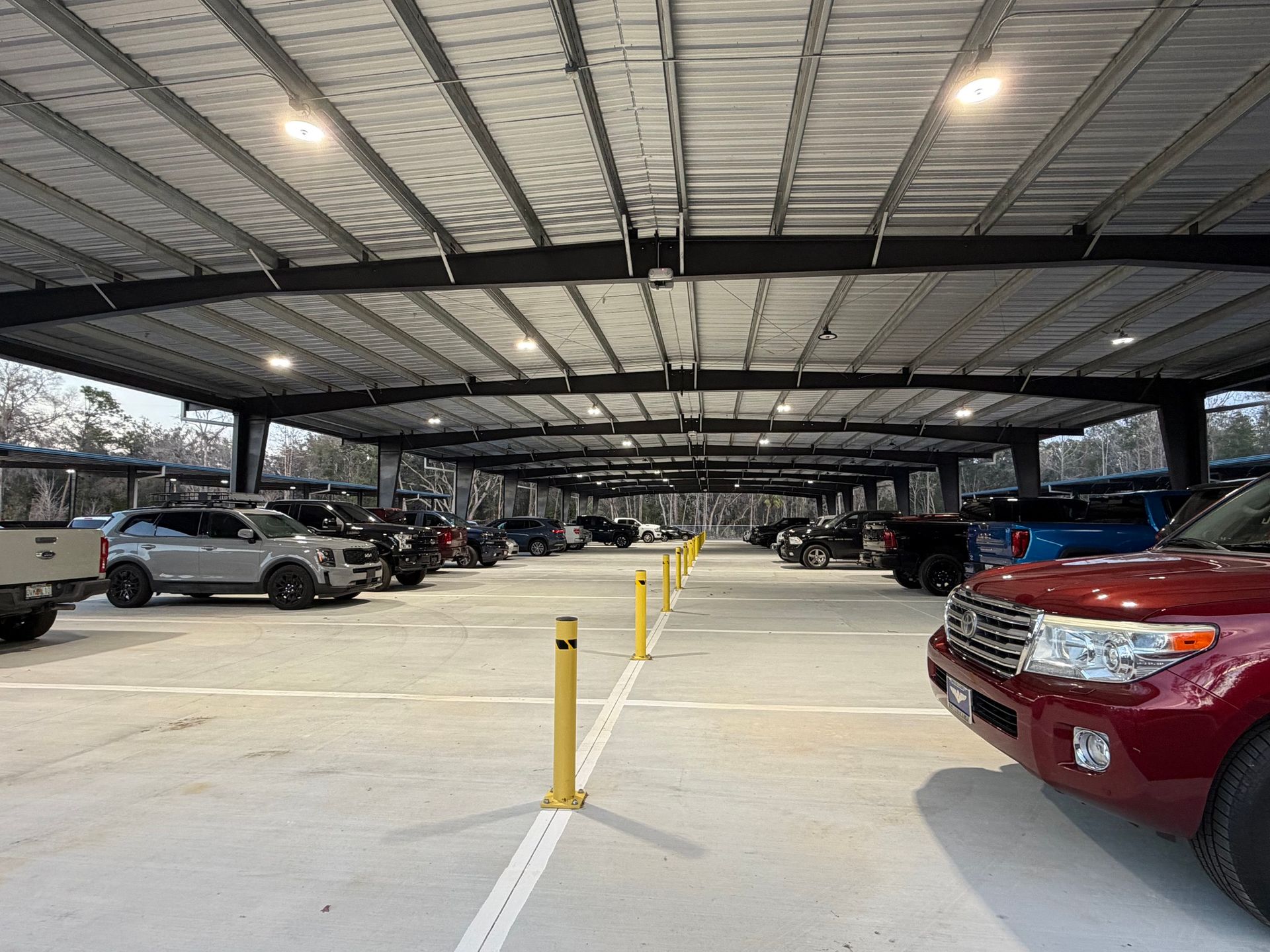 A covered parking structure with several vehicles parked, featuring overhead lighting and yellow concrete bollards.