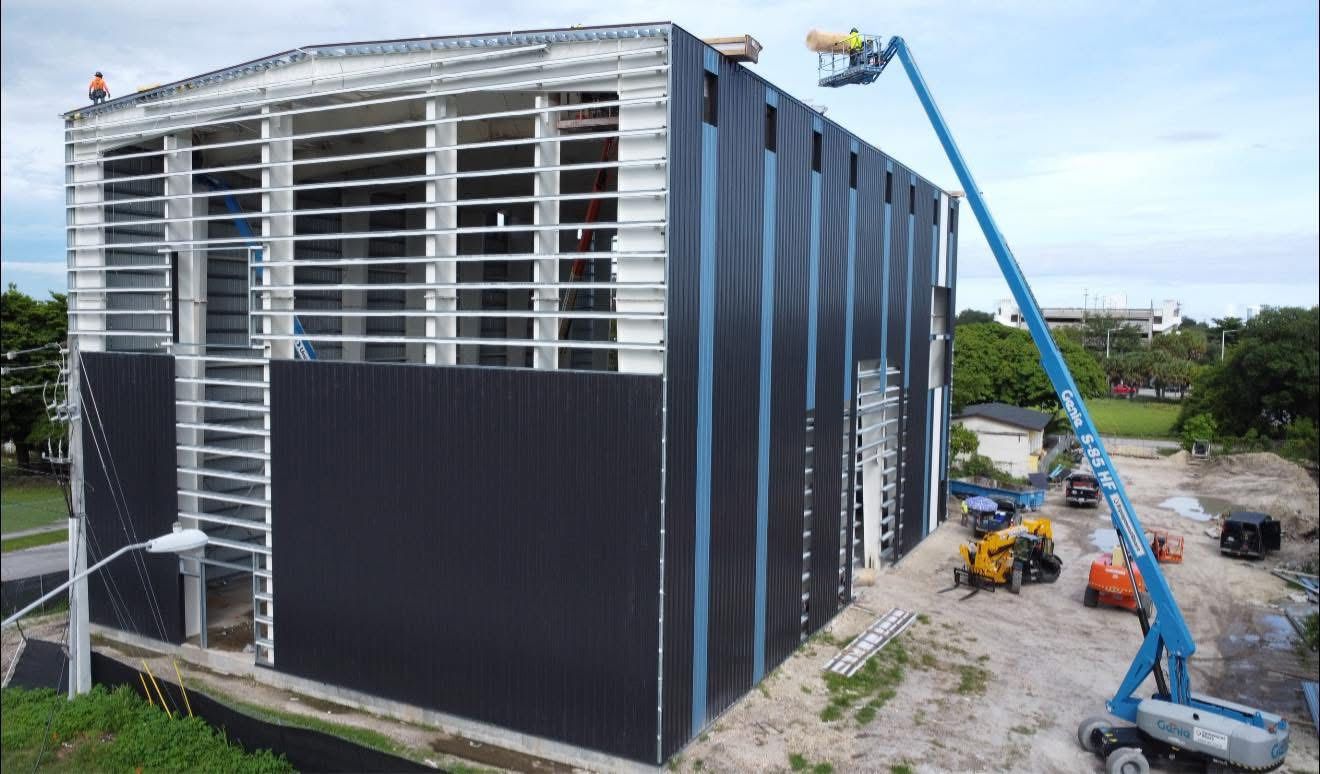 A construction site with a partially metal-clad building and a blue boom lift reaching the roof under a cloudy sky.