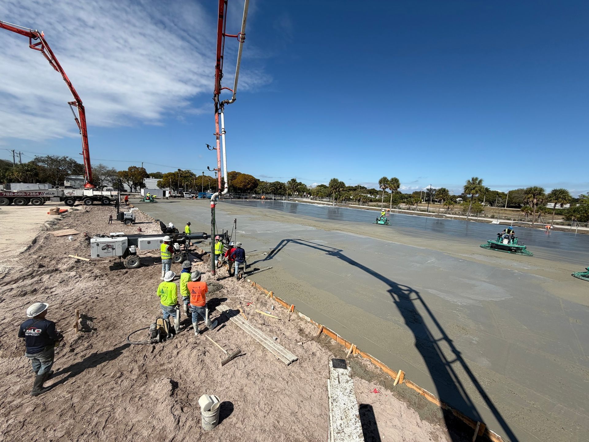 Construction workers oversee concrete being poured across a large, level site by two long boom pumps under a sunny sky.