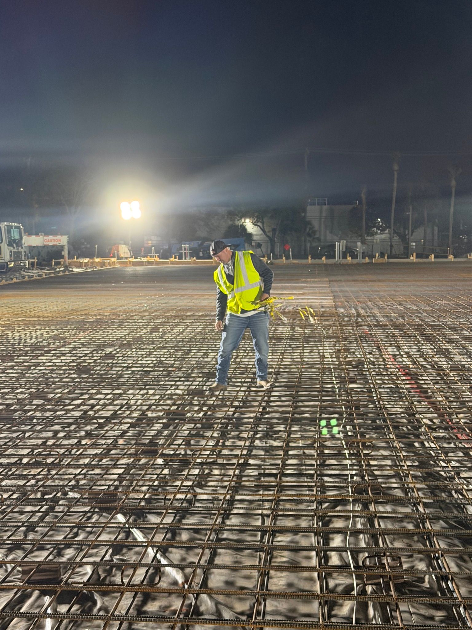 A worker in a high-visibility vest stands on a rebar grid at a brightly lit outdoor construction site at night.