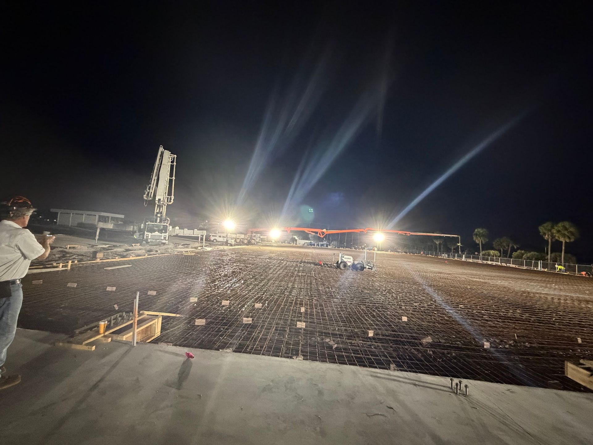 A person watches a construction site illuminated by bright floodlights at night, with a concrete pump in the background.