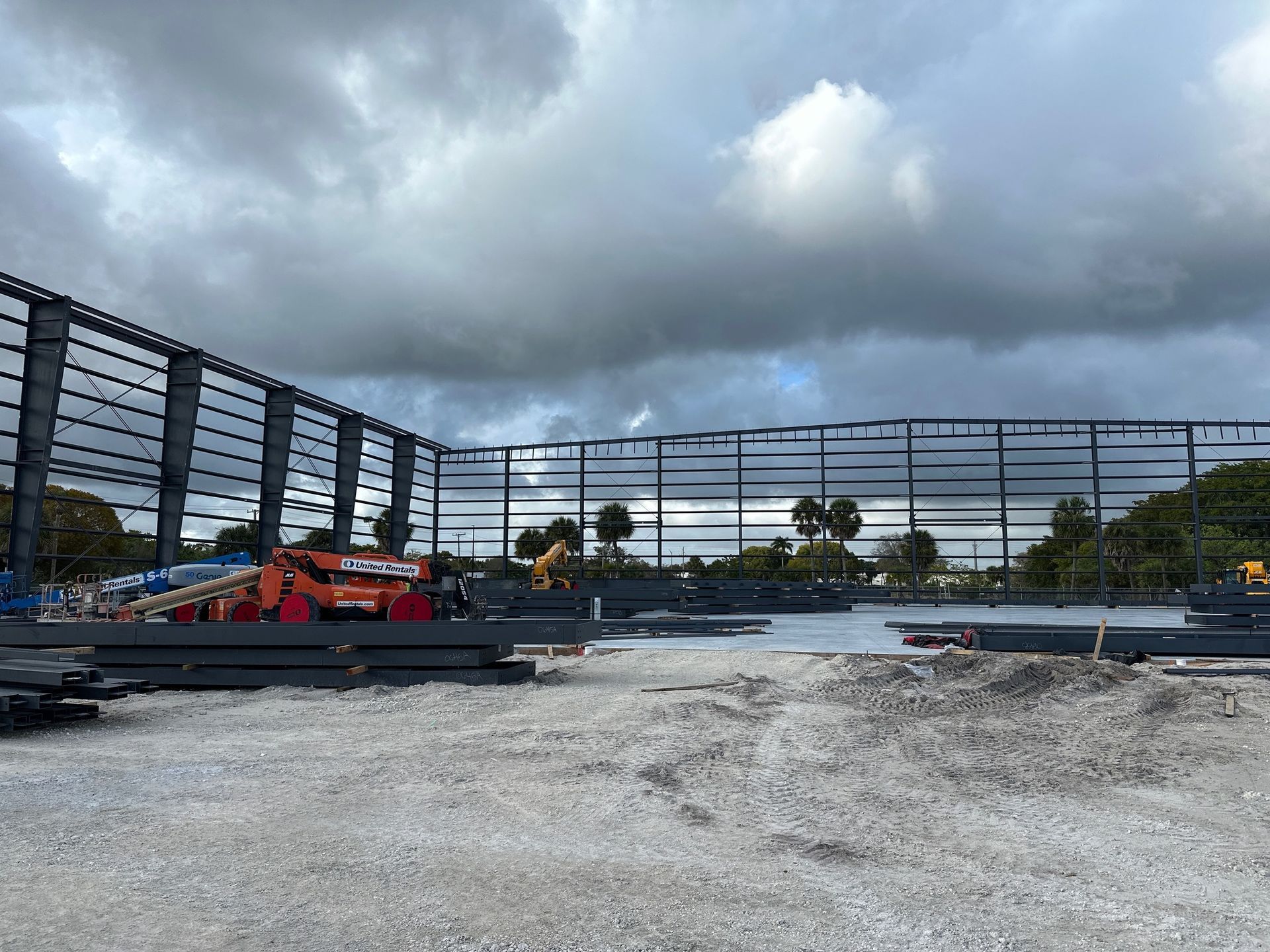 Steel framing under construction at a worksite with heavy equipment, piles of building materials, and a cloudy sky.