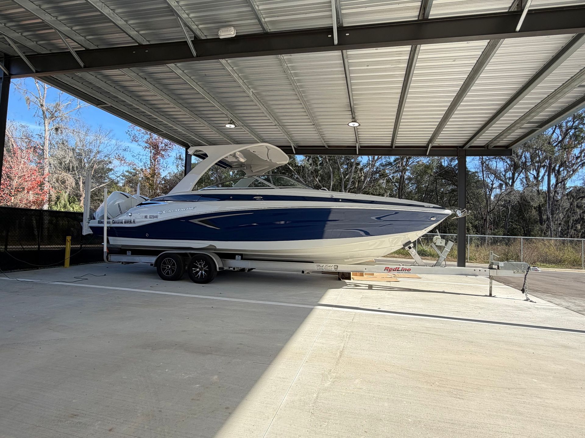 A blue and white motorboat on a trailer parked under a metal-roofed shelter outdoors on a sunny day.