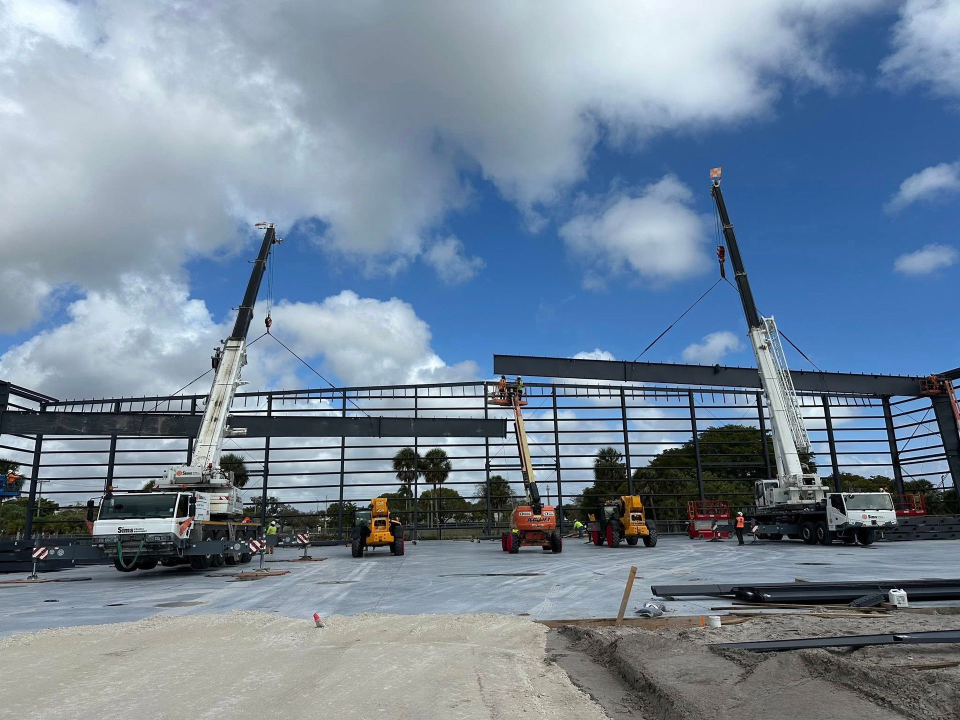 Two large cranes lift a long steel beam into position for the construction of a large metal building under a blue sky.