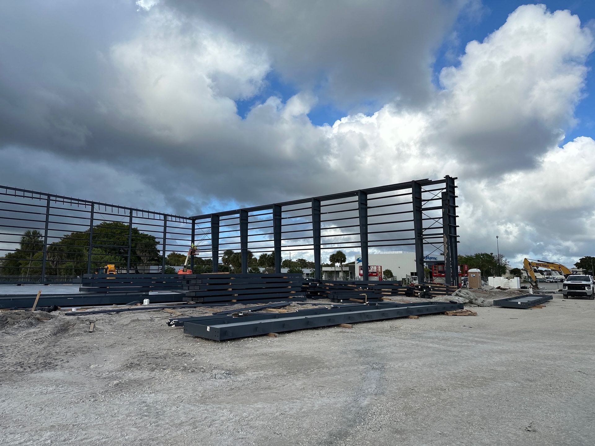 Construction site showing a large, dark steel frame for a new building against a bright, cloudy blue sky.