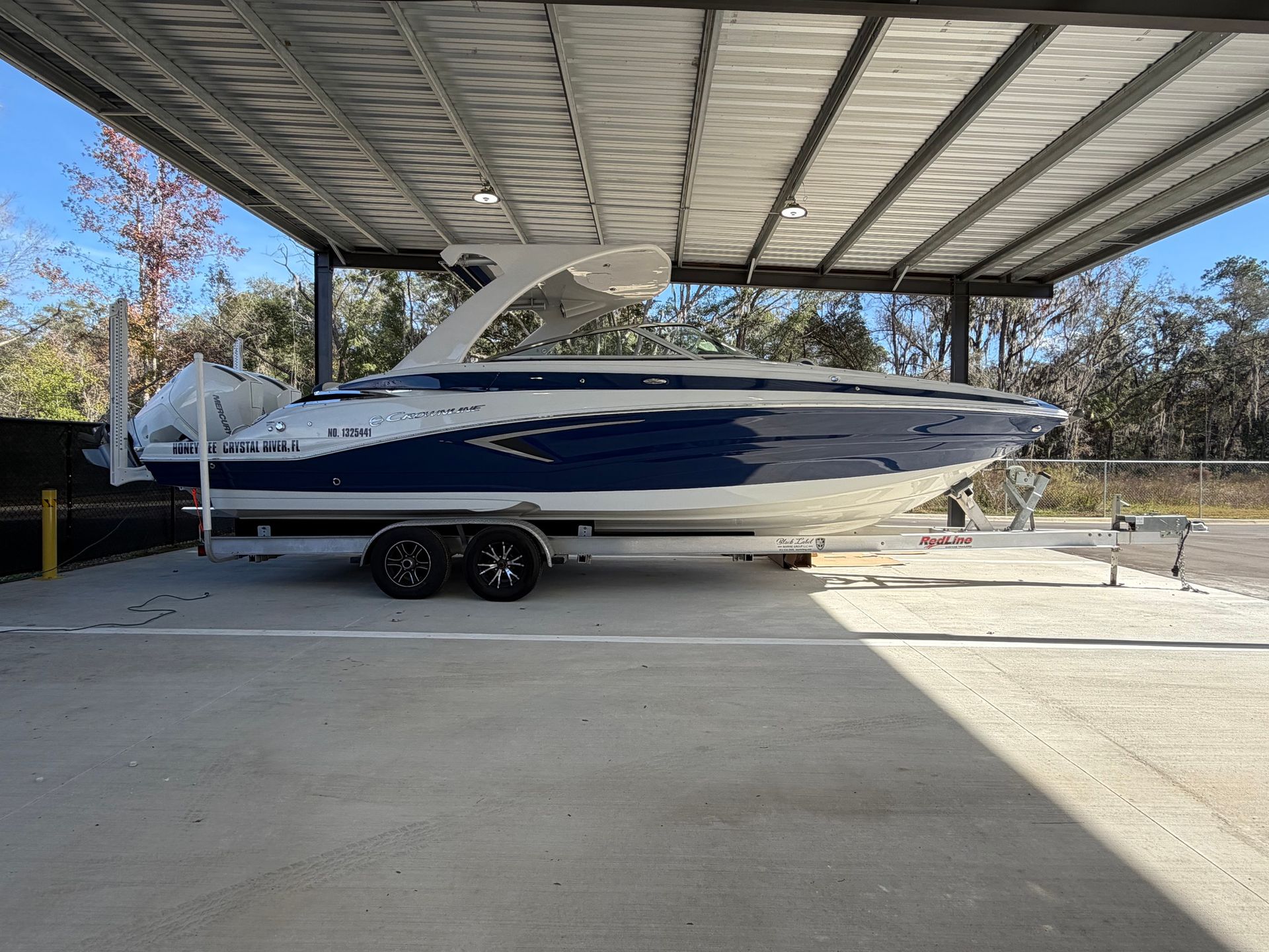 A blue and white motorboat on a trailer parked under a metal canopy structure on a concrete lot.