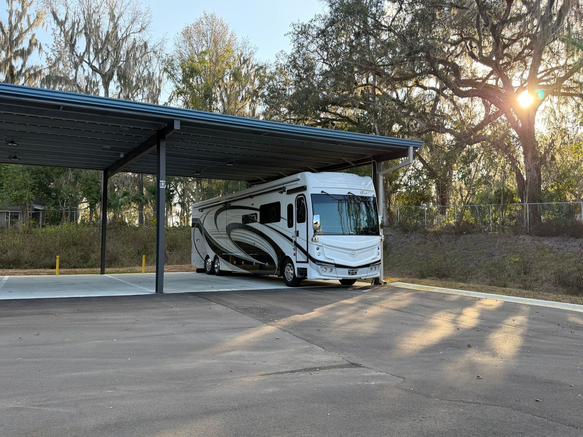A white and black recreational vehicle is parked under a metal-roofed carport surrounded by trees at sunset.