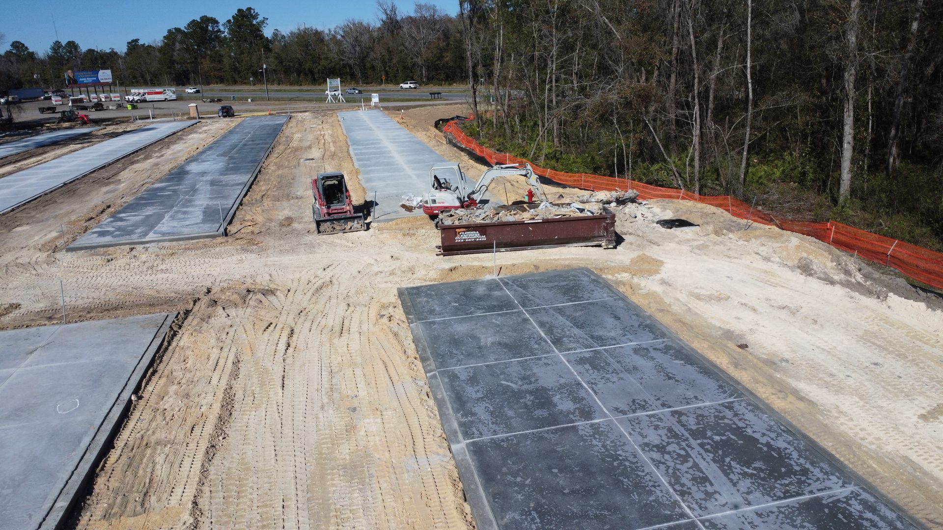 An aerial view of a construction site with several rectangular concrete slabs laid out on sandy ground near trees.