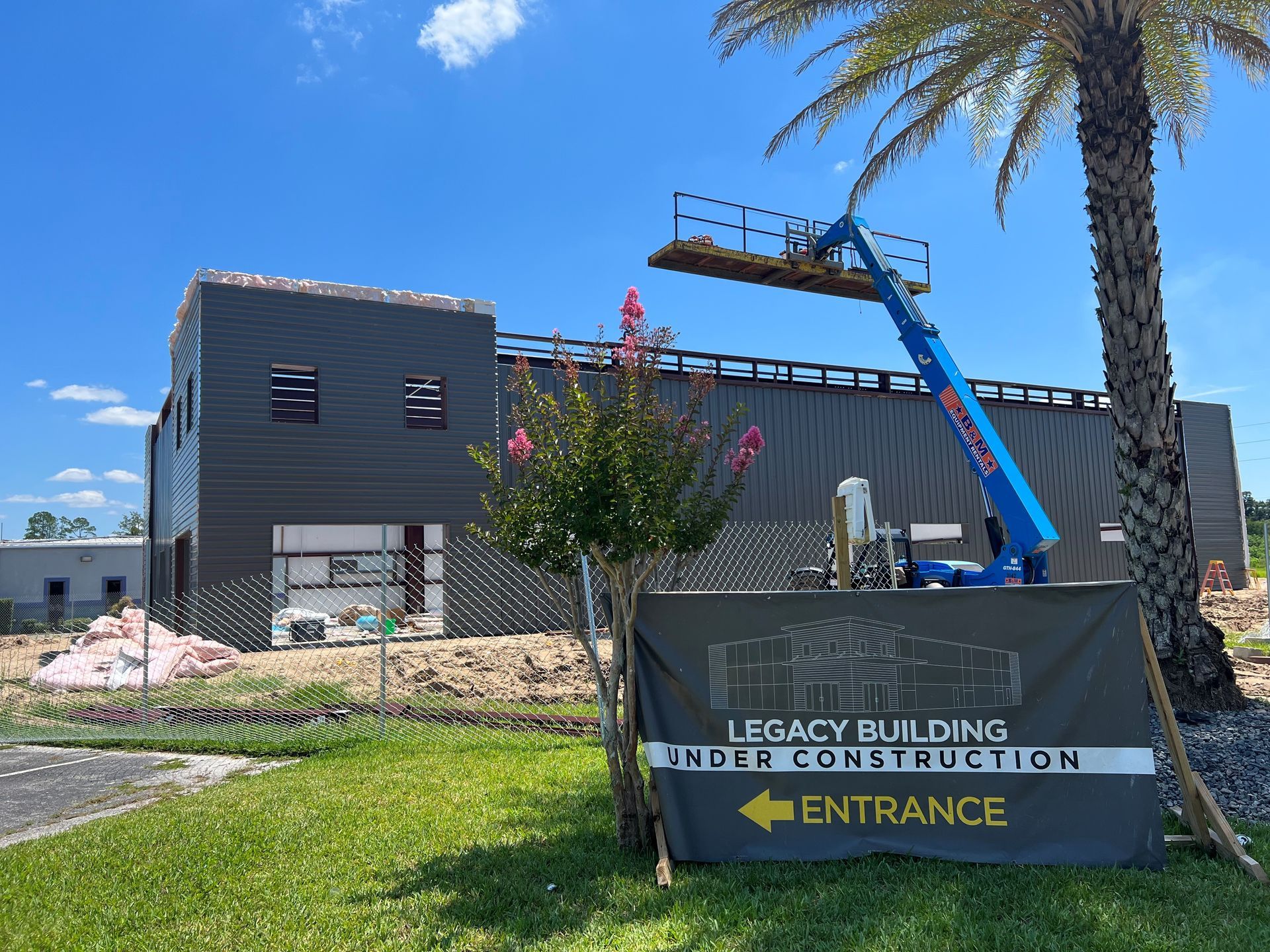 A construction site featuring a dark-paneled building, a blue construction lift, and a sign directing to the entrance.