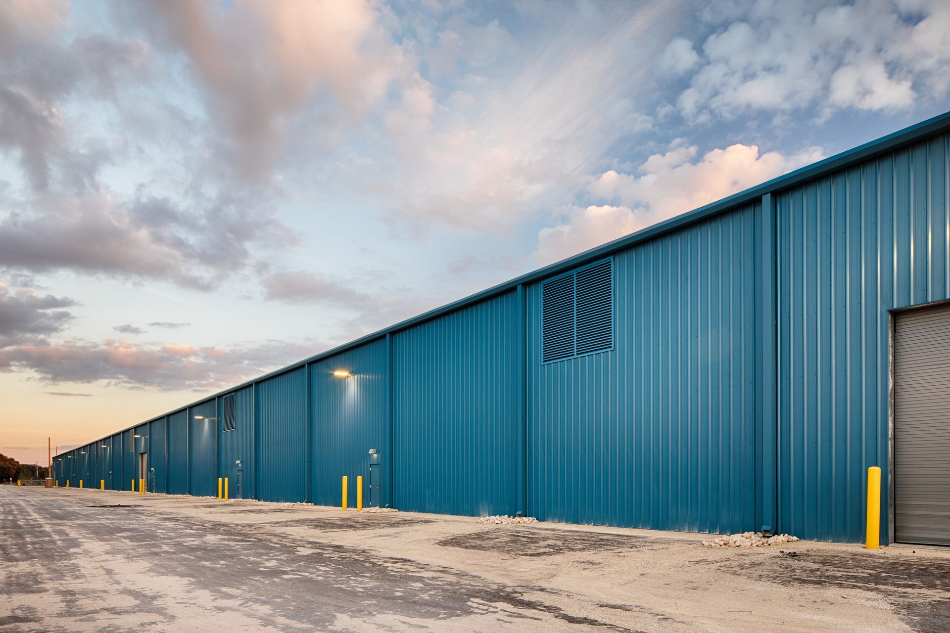 A long, blue corrugated metal warehouse building under a cloudy sky at sunset, with a dirt lot in the foreground.