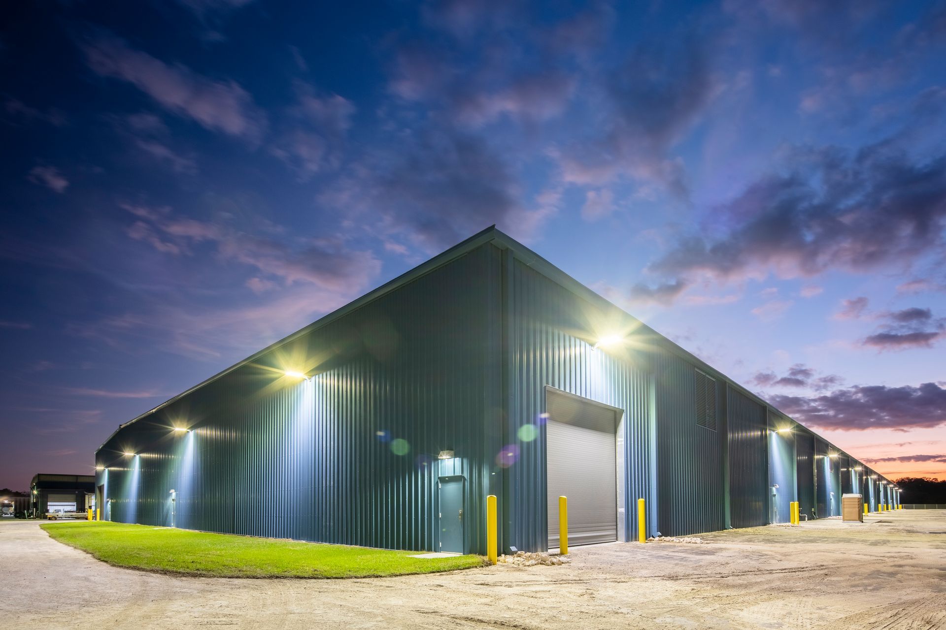 A dark blue industrial warehouse with metal siding and roll-up doors, illuminated by exterior lights at twilight.
