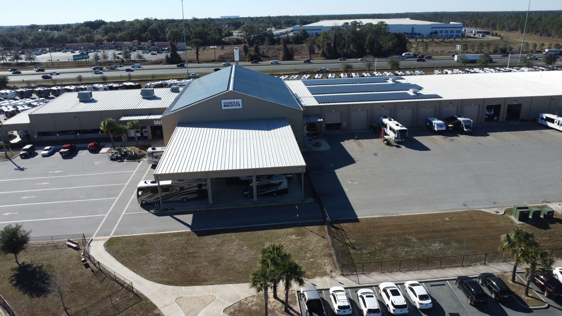 Aerial view of a commercial dealership building with a white peaked awning, parking lot, and nearby highway.