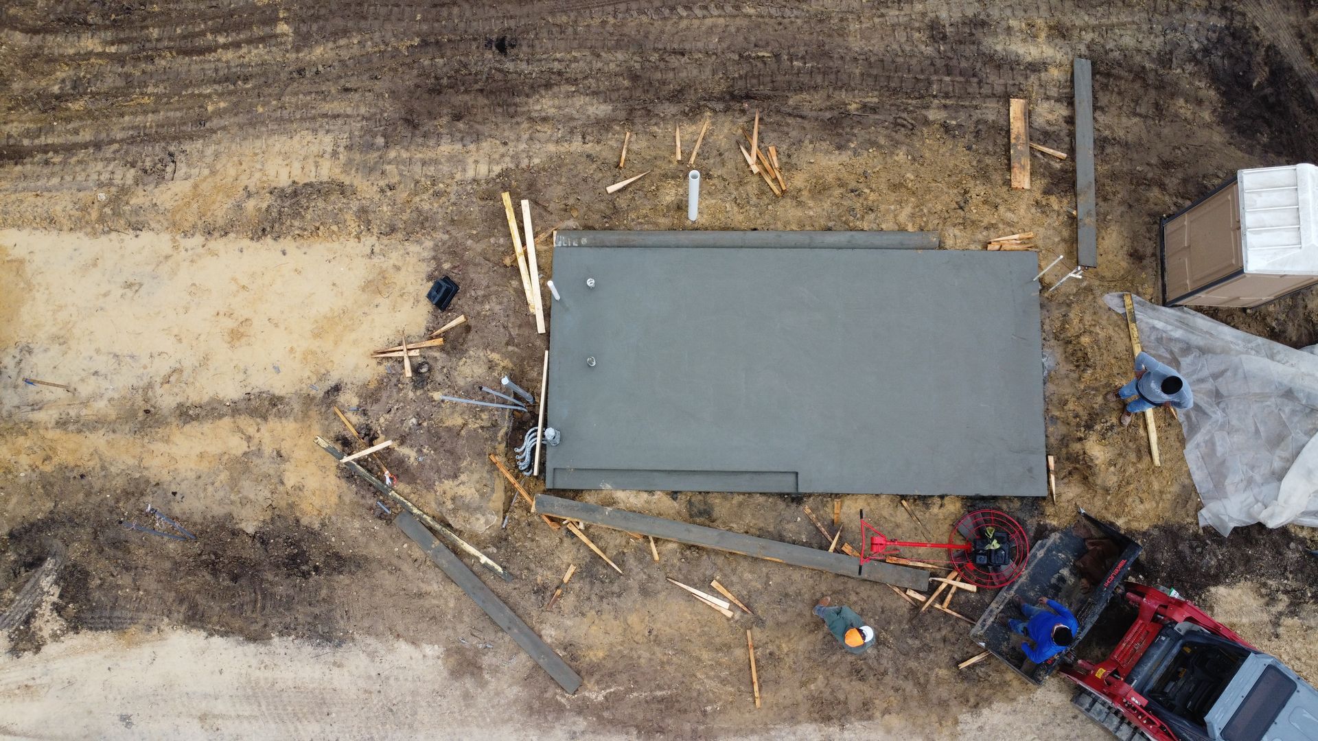 Aerial view of workers at a muddy construction site pouring a rectangular concrete slab foundation.
