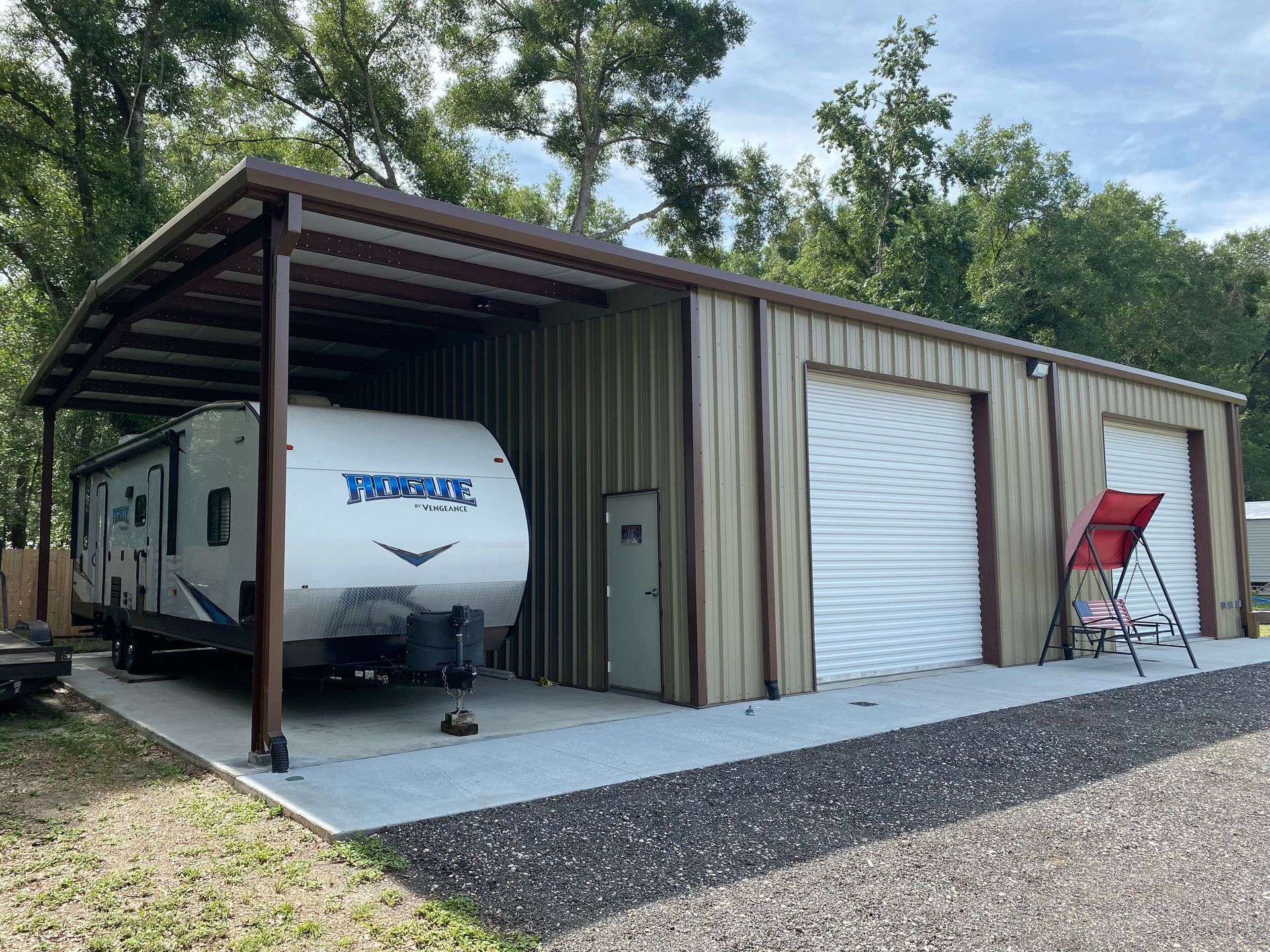 A tan metal building with an attached carport sheltering a white travel trailer, set on a concrete pad with gravel.