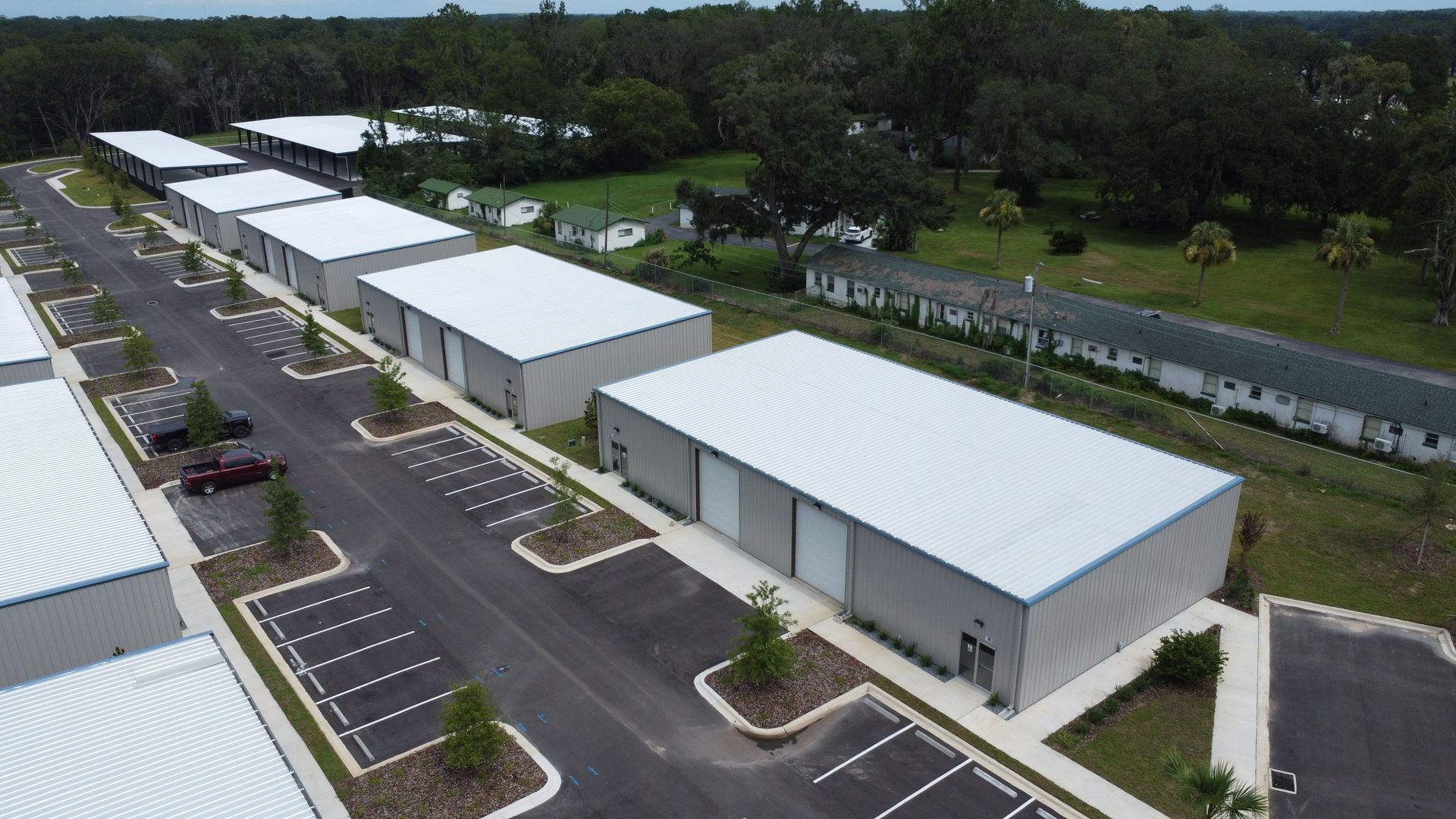 Aerial view of a complex of industrial warehouse buildings with white roofs and paved parking lots surrounded by trees.