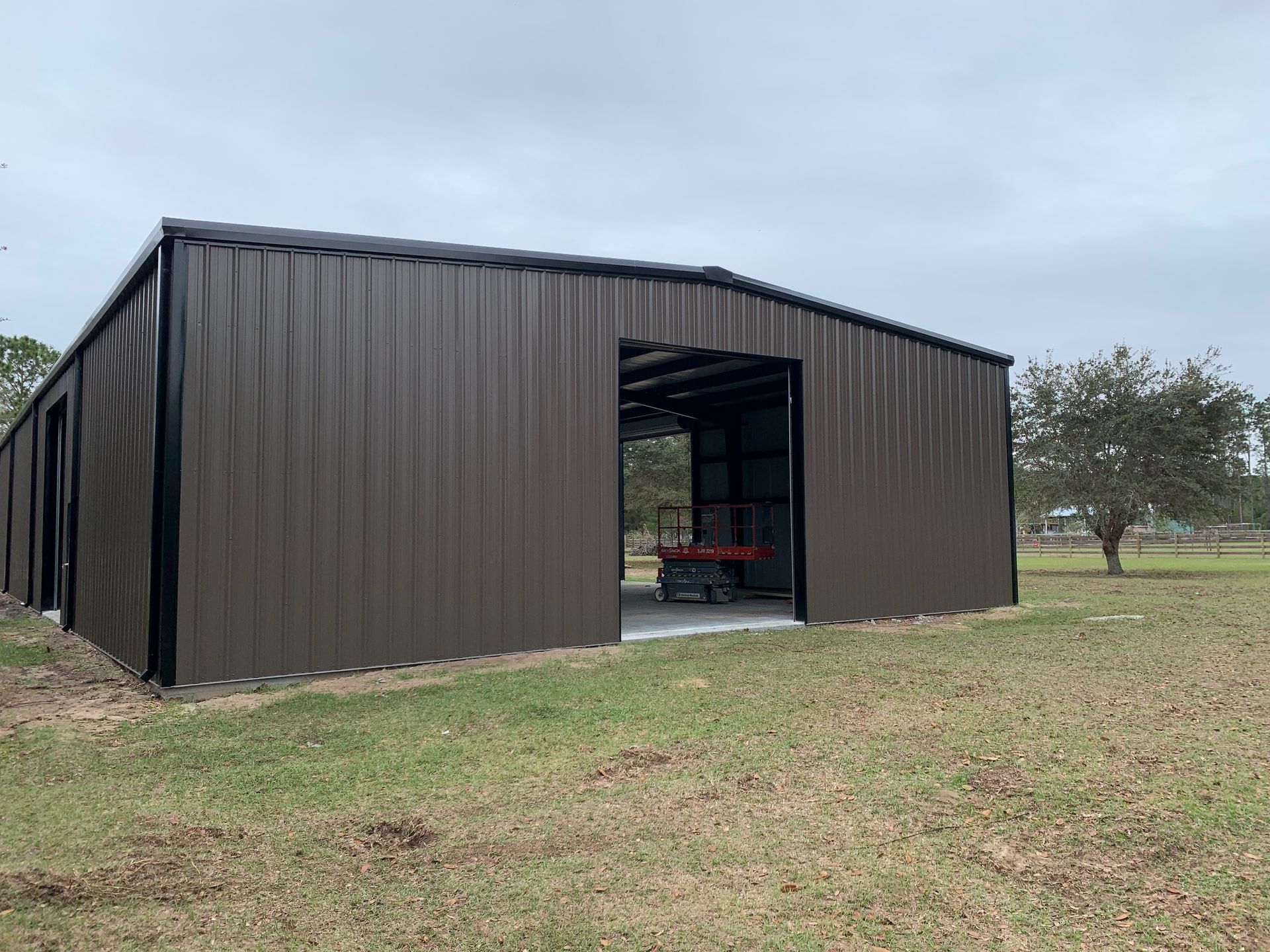 A dark brown, metal-sided barn with an open doorway stands on a grassy field under a cloudy sky.