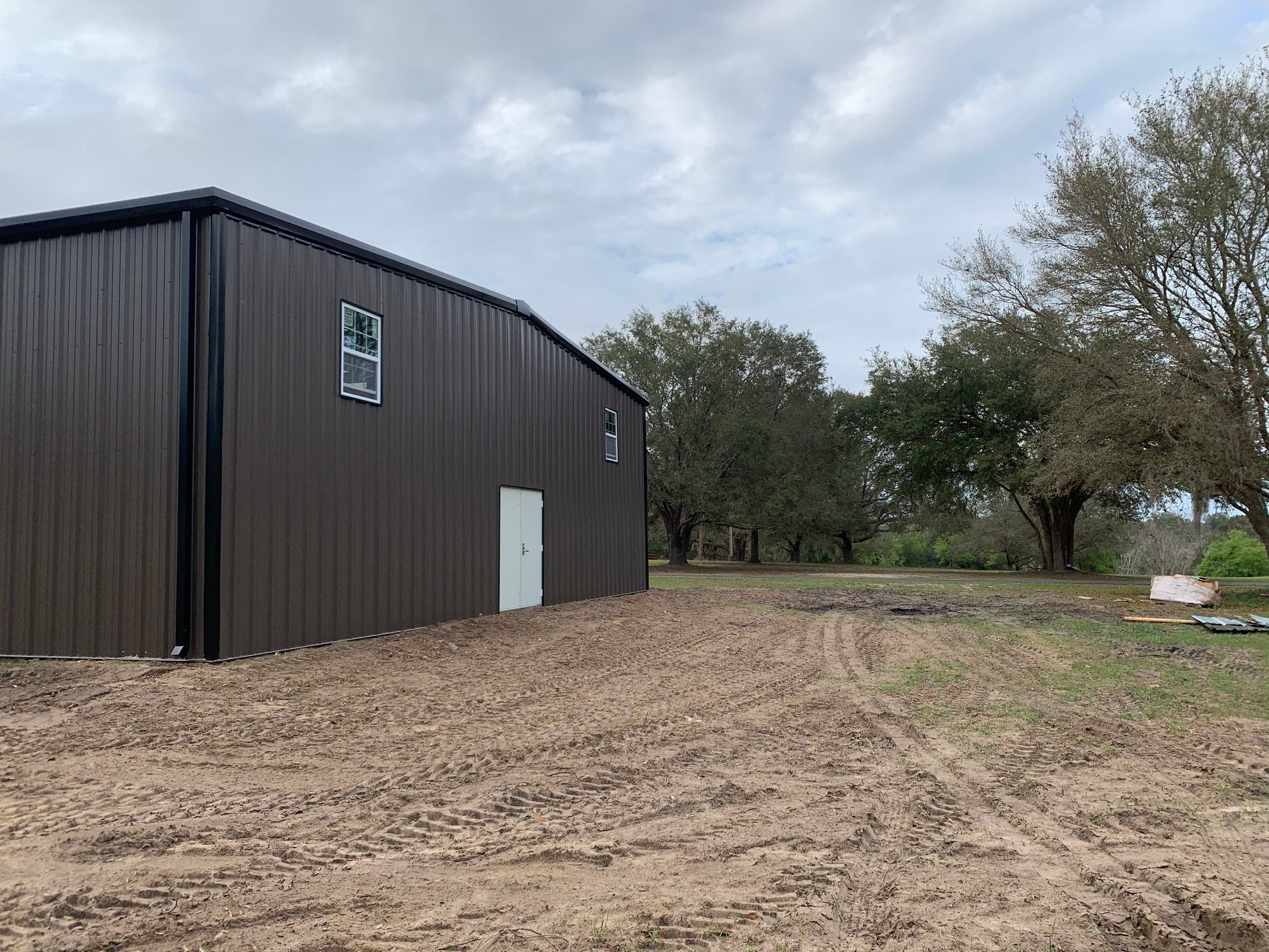 A dark-brown metal warehouse stands on a dirt lot with mature oak trees under a cloudy sky.