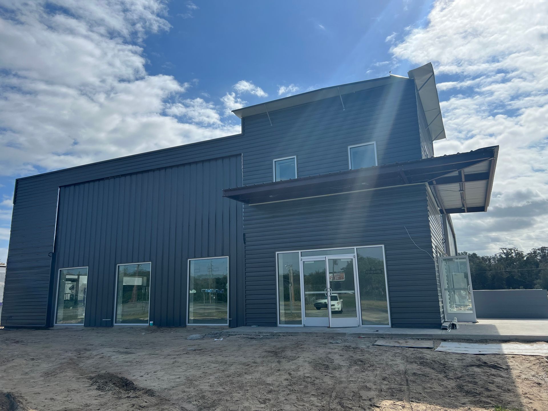 A newly constructed building with dark metal siding, glass doors, and windows, set against a bright, cloudy sky.