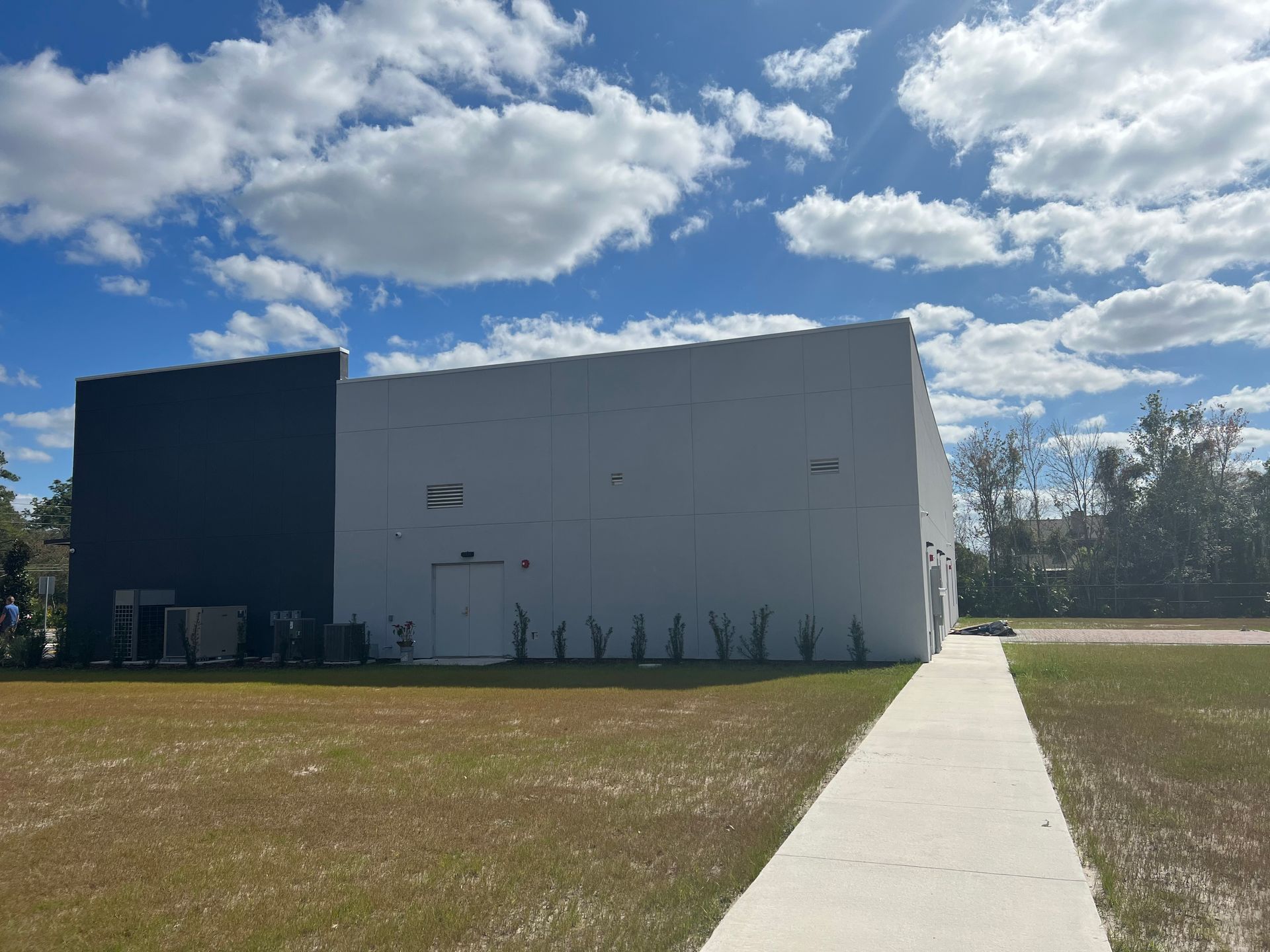 A two-tone white and dark blue building stands under a bright, partly cloudy sky next to a sidewalk and grassy field.