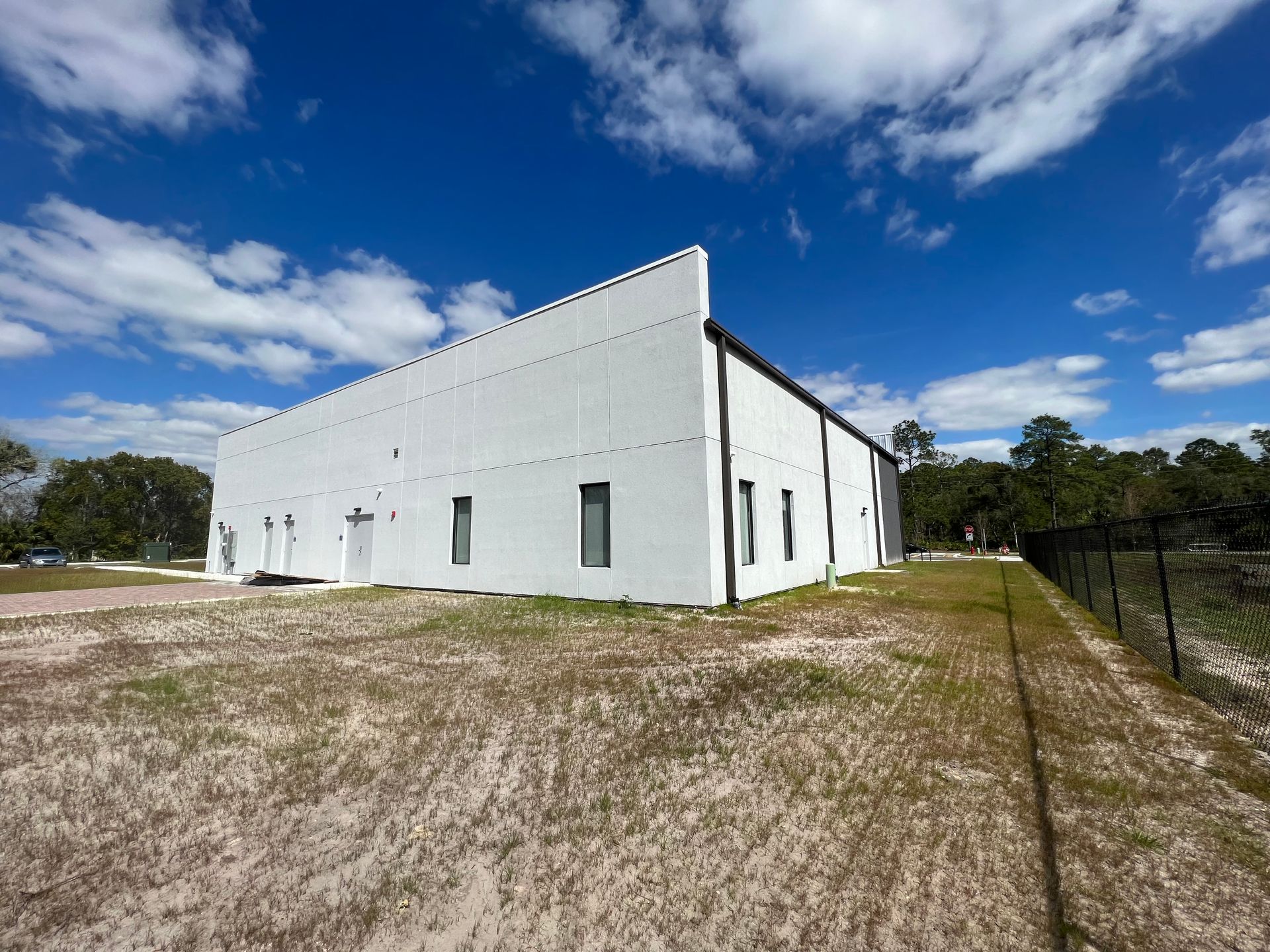 A white, rectangular commercial building sits in an open, grassy lot under a blue sky with scattered clouds.