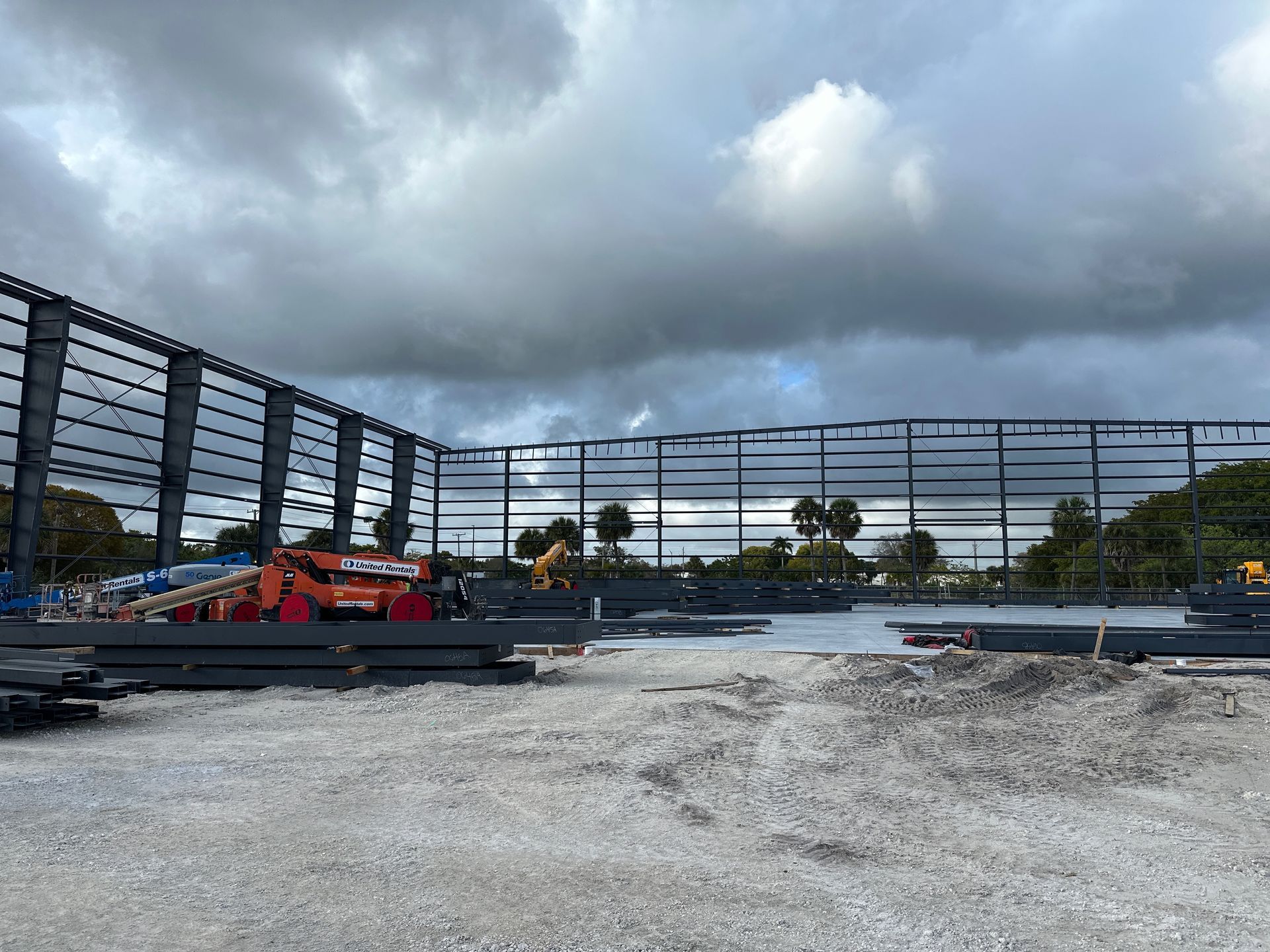A steel frame skeleton of a large building under construction on a gravel lot under a cloudy sky.