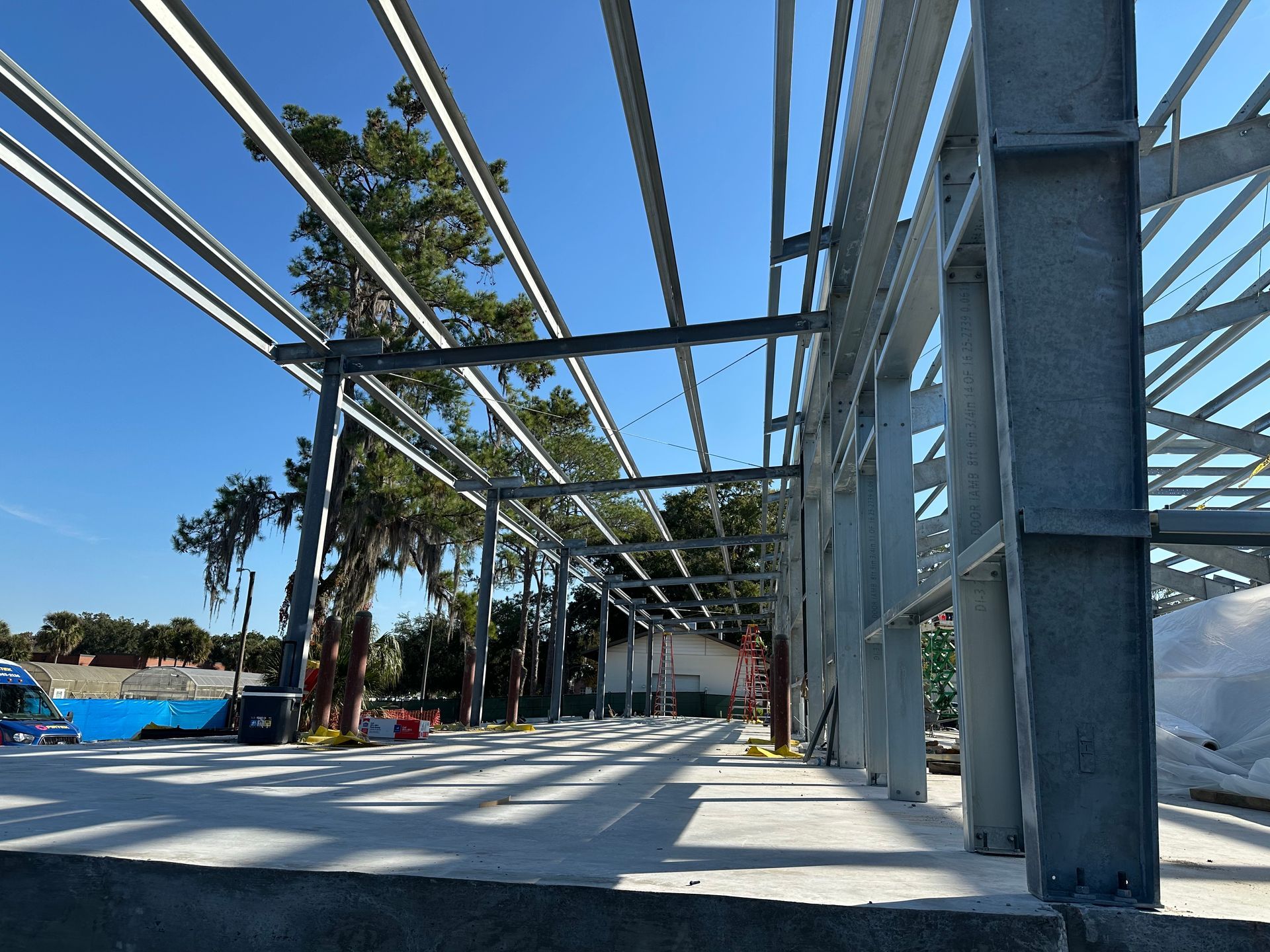 A construction site with an exposed steel frame for a building, concrete slab foundation, and trees against a blue sky.
