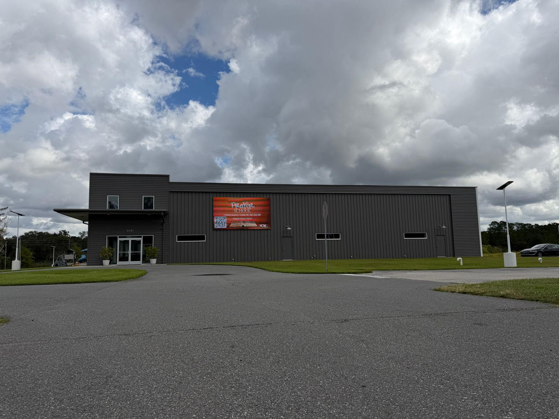 A modern gray industrial building with a prominent red sign and glass entrance under a cloudy sky.