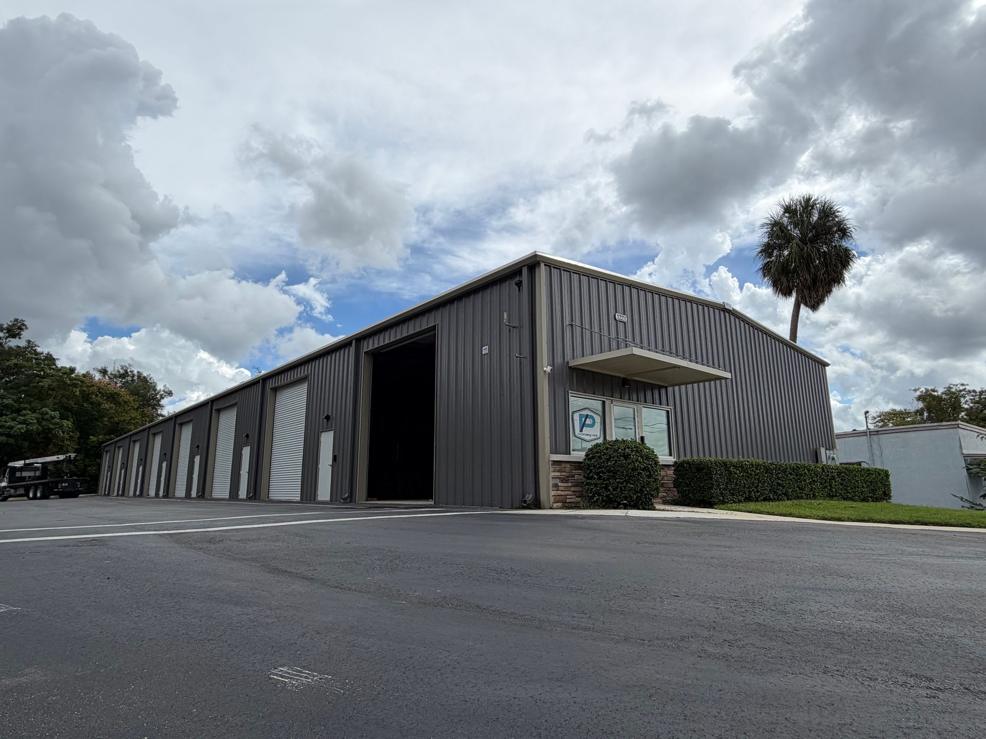 A long, grey metal commercial building with multiple roll-up garage doors under a cloudy sky with a single palm tree.