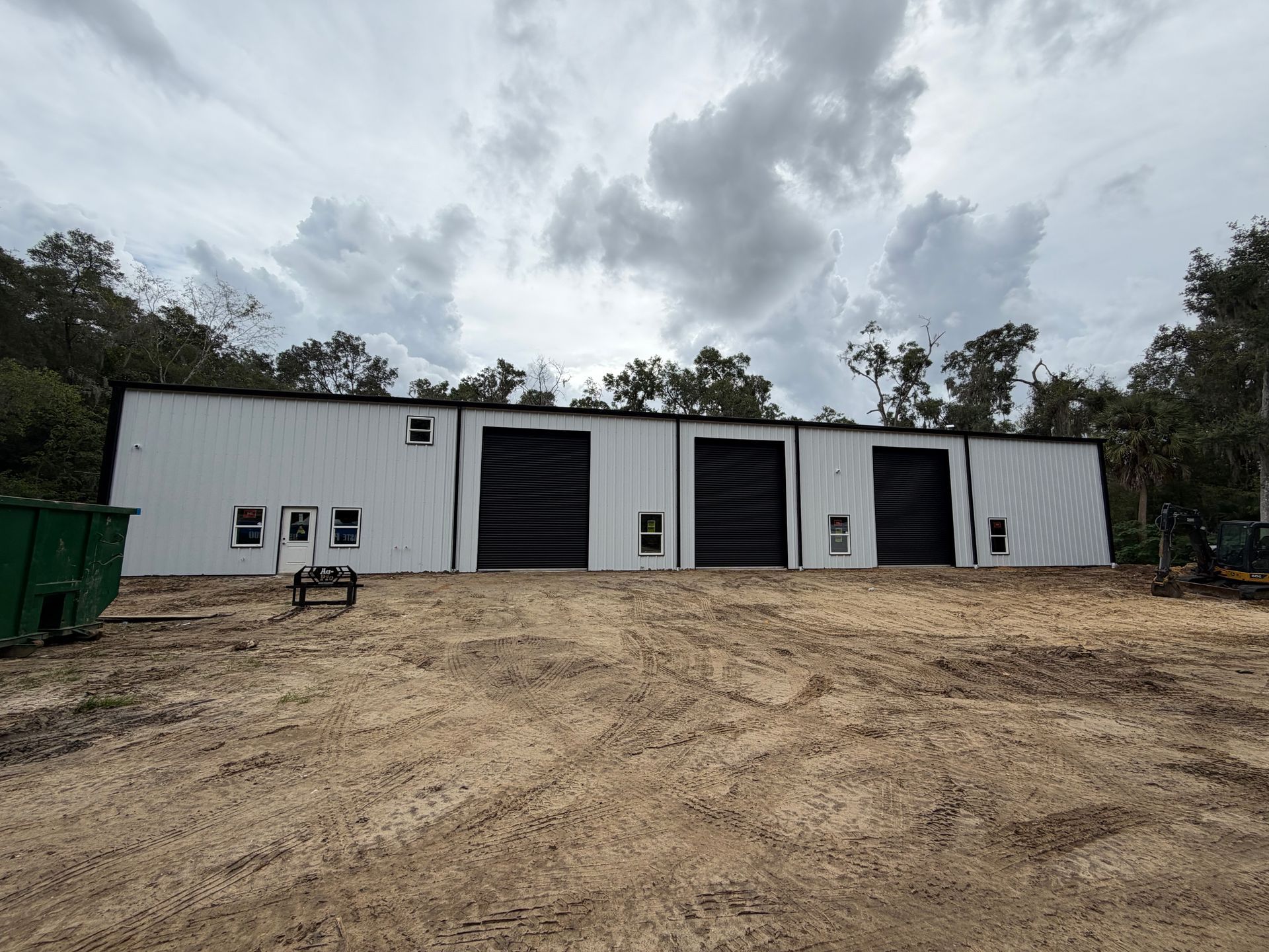 A white metal warehouse building with three large black roll-up doors, set on a dirt lot with a green dumpster nearby.