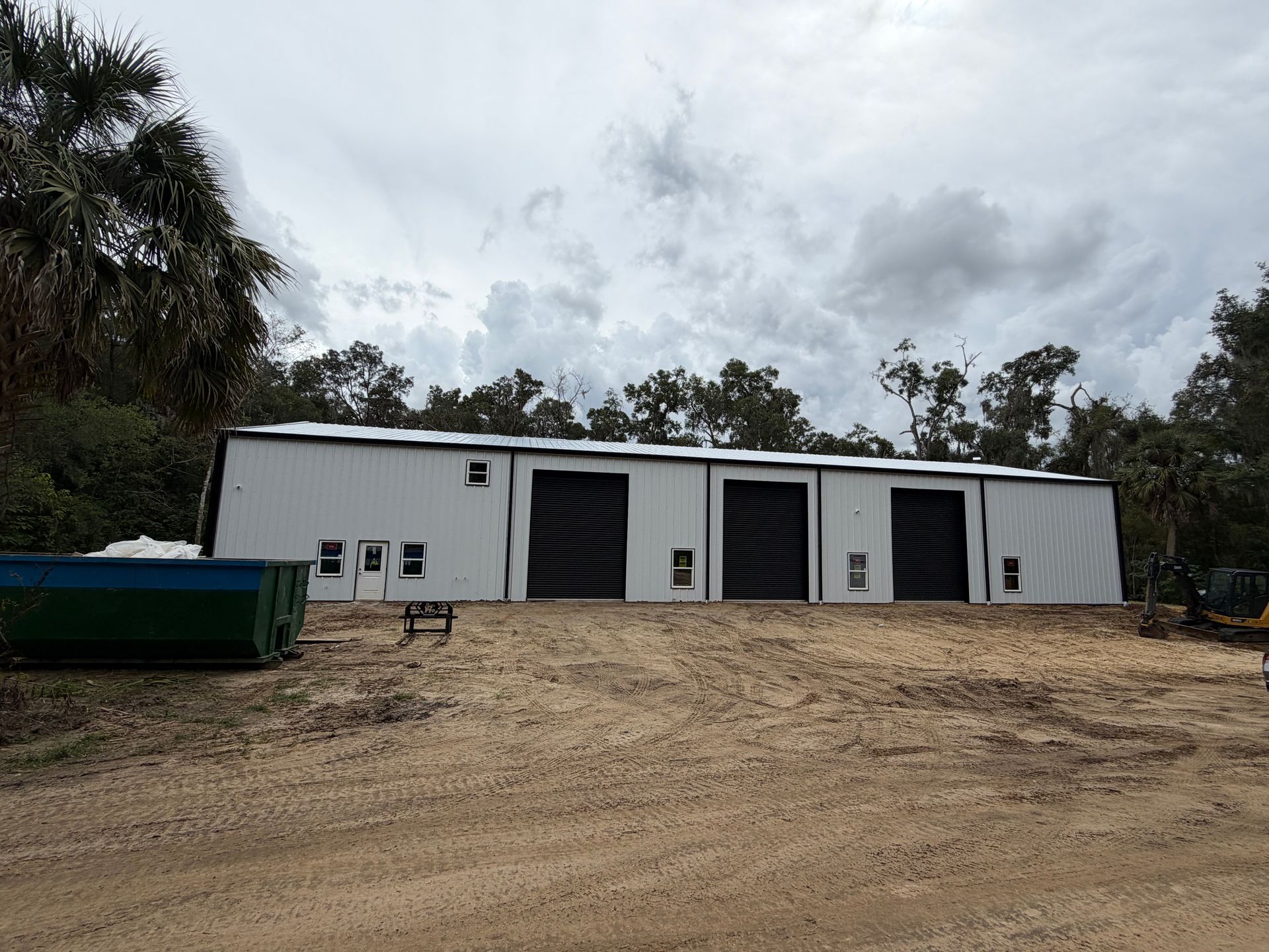 A large white industrial metal building with three black rolling doors, set on a dirt lot with a dumpster and trees.